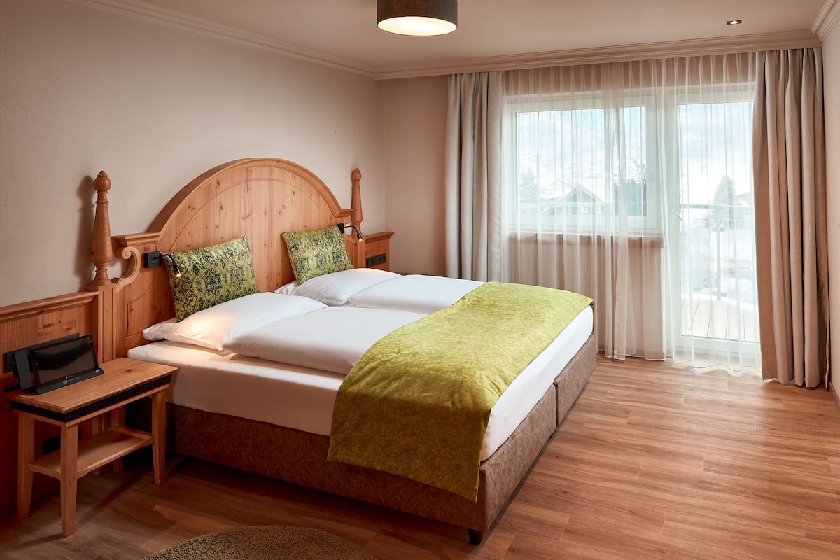 A neatly made bed with a wooden headboard, a bedside table with a lamp, and a view of the outdoors through sheer curtains.