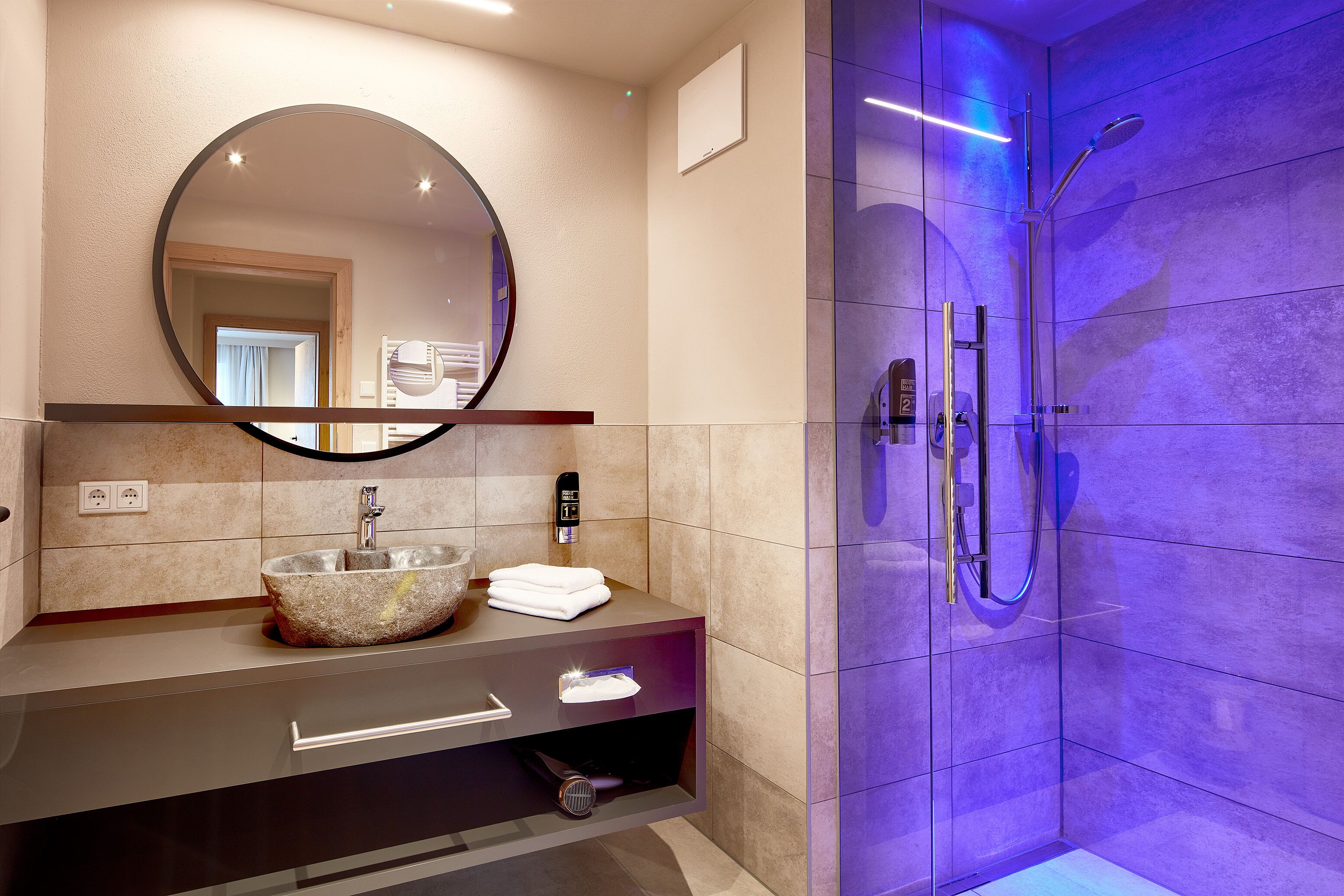 A modern bathroom with a round mirror, a stone sink, a built-in towel rack, and a walk-in shower with a handheld showerhead.