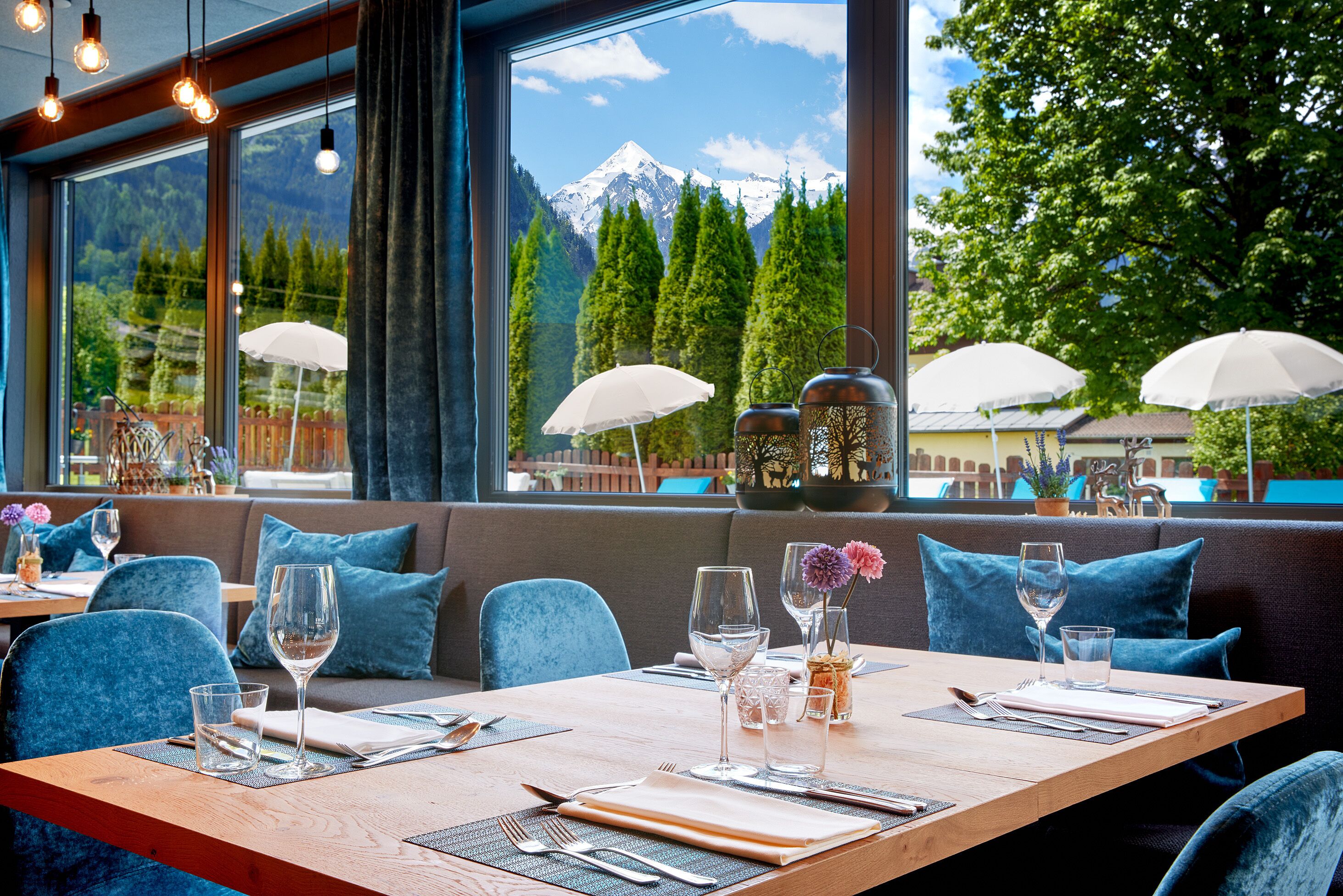 Un restaurant avec une table en bois dressée pour le repas, une vue sur les montagnes à travers de grandes fenêtres et des chaises recouvertes d’un tissu bleu.