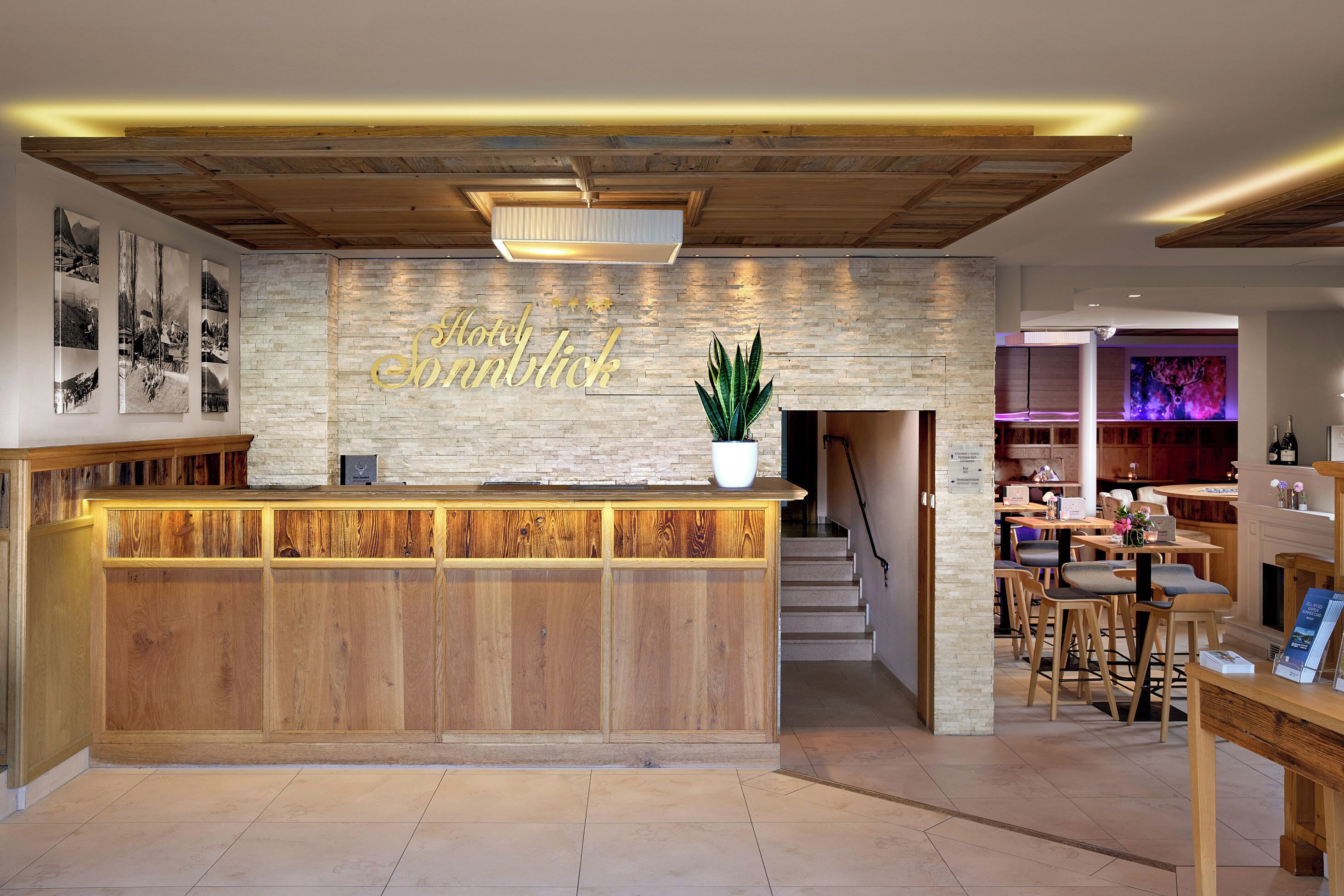 A hotel reception area with a wooden reception desk, a large potted plant, and a wall with the hotel's name. There are stairs leading to a dining area with tables and chairs.
