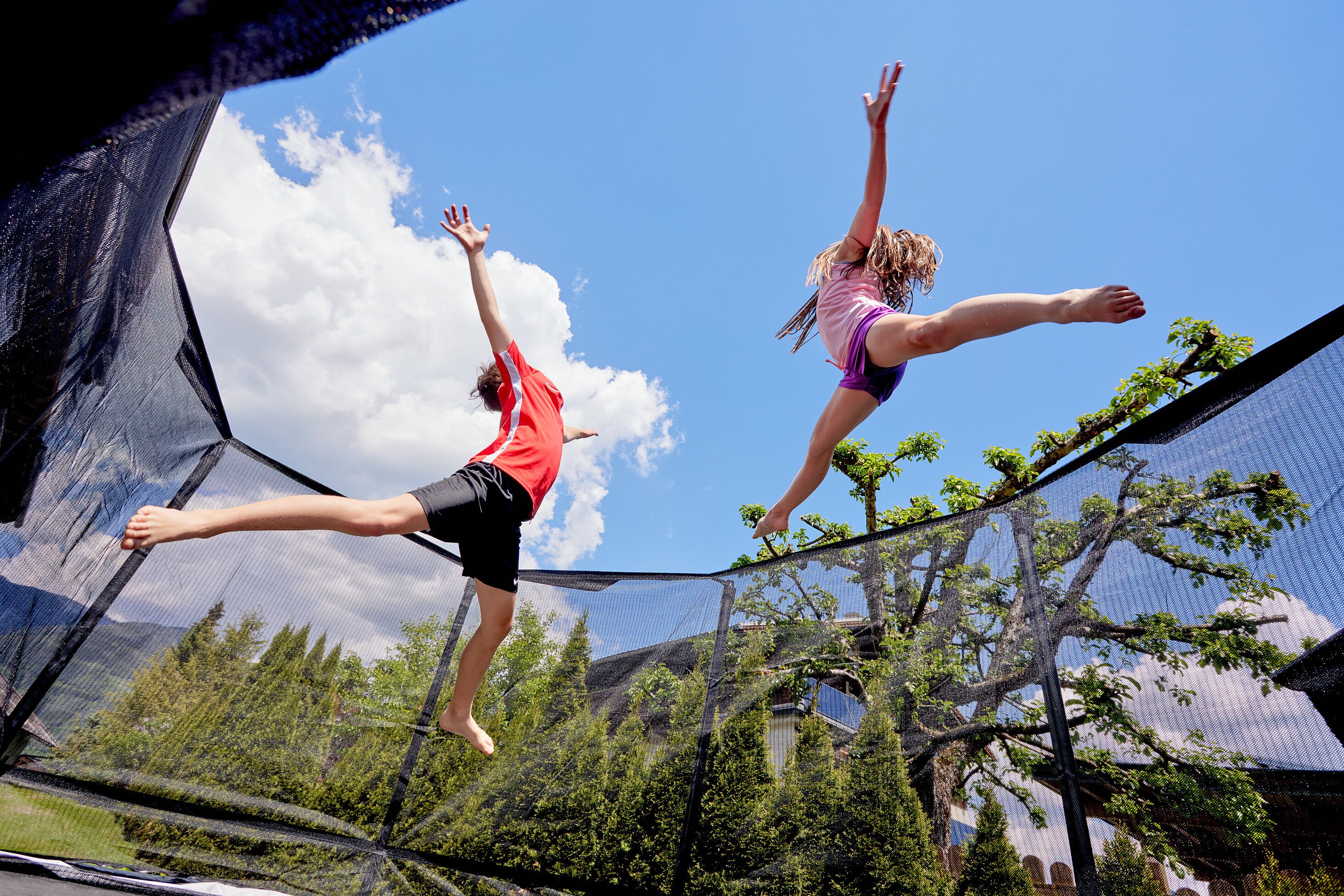 Two people jumping on a trampoline with a clear sky and trees in the background.