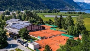 A tennis court surrounded by mountains, a parking area, and buildings with solar panels. - Hotel Sonnblick (Kaprun)
