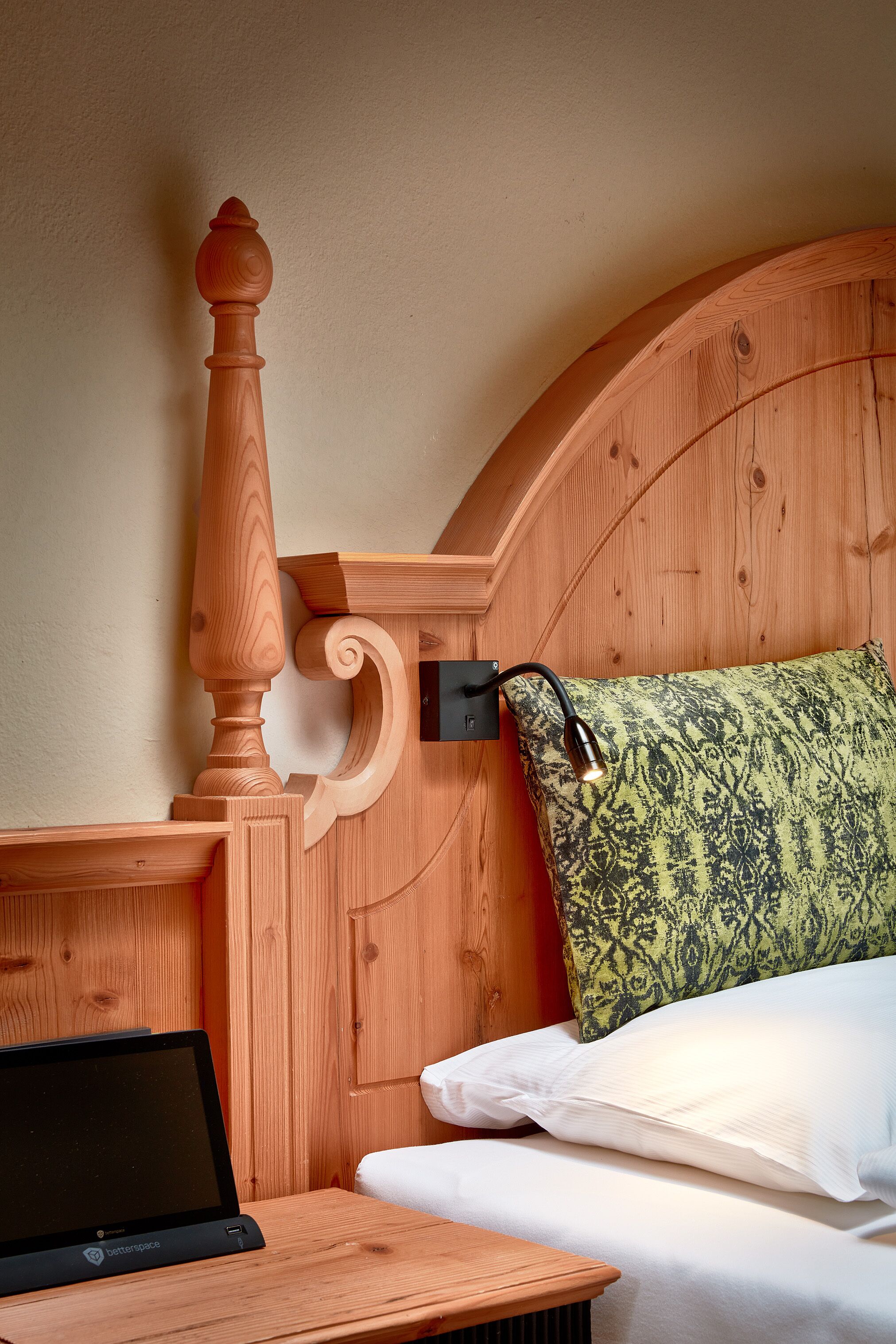 Cabecera de madera con lámpara integrada, una laptop sobre una mesita de noche de madera y una cama con almohada estampada.