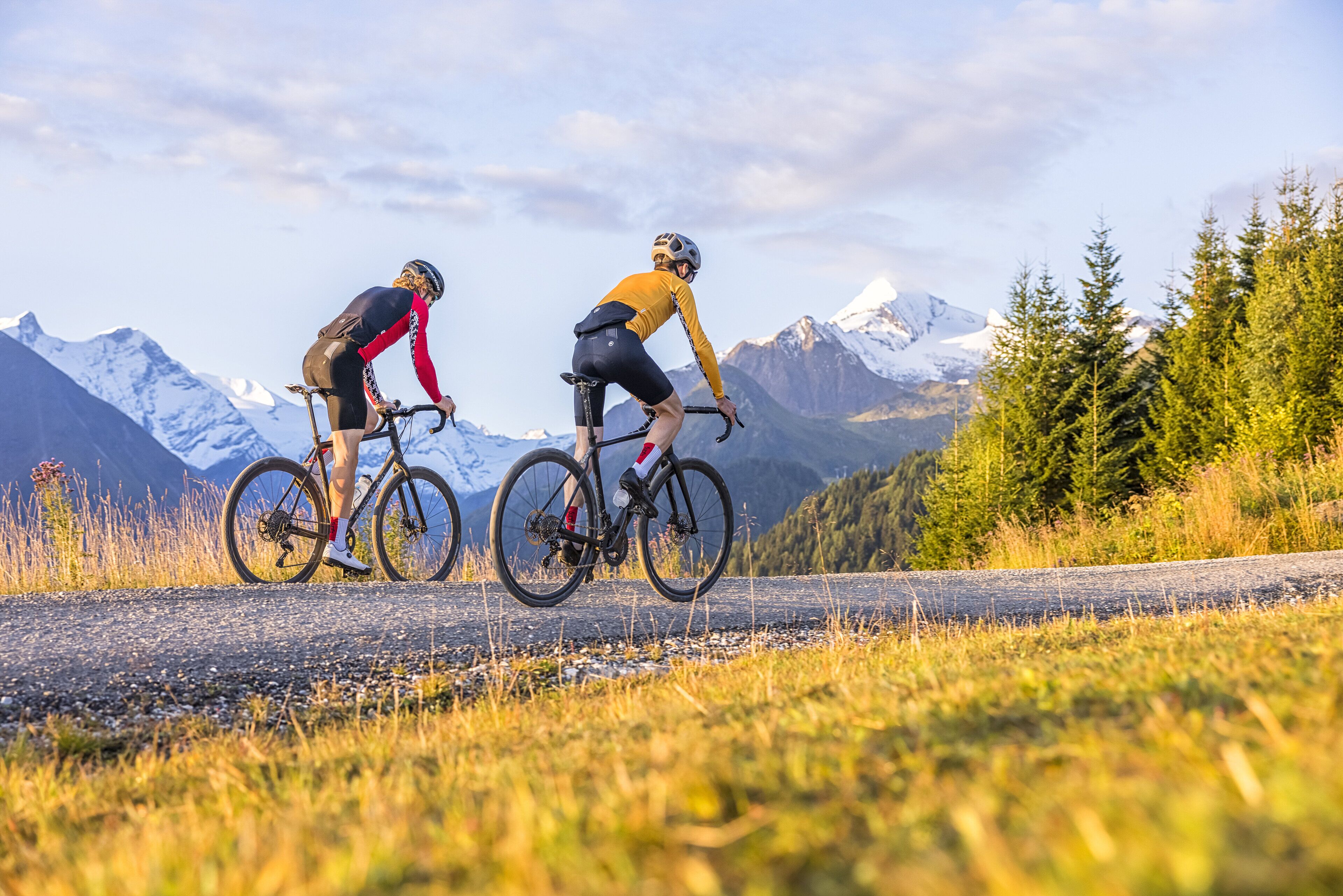 Two cyclists on a gravel road with a mountainous backdrop.