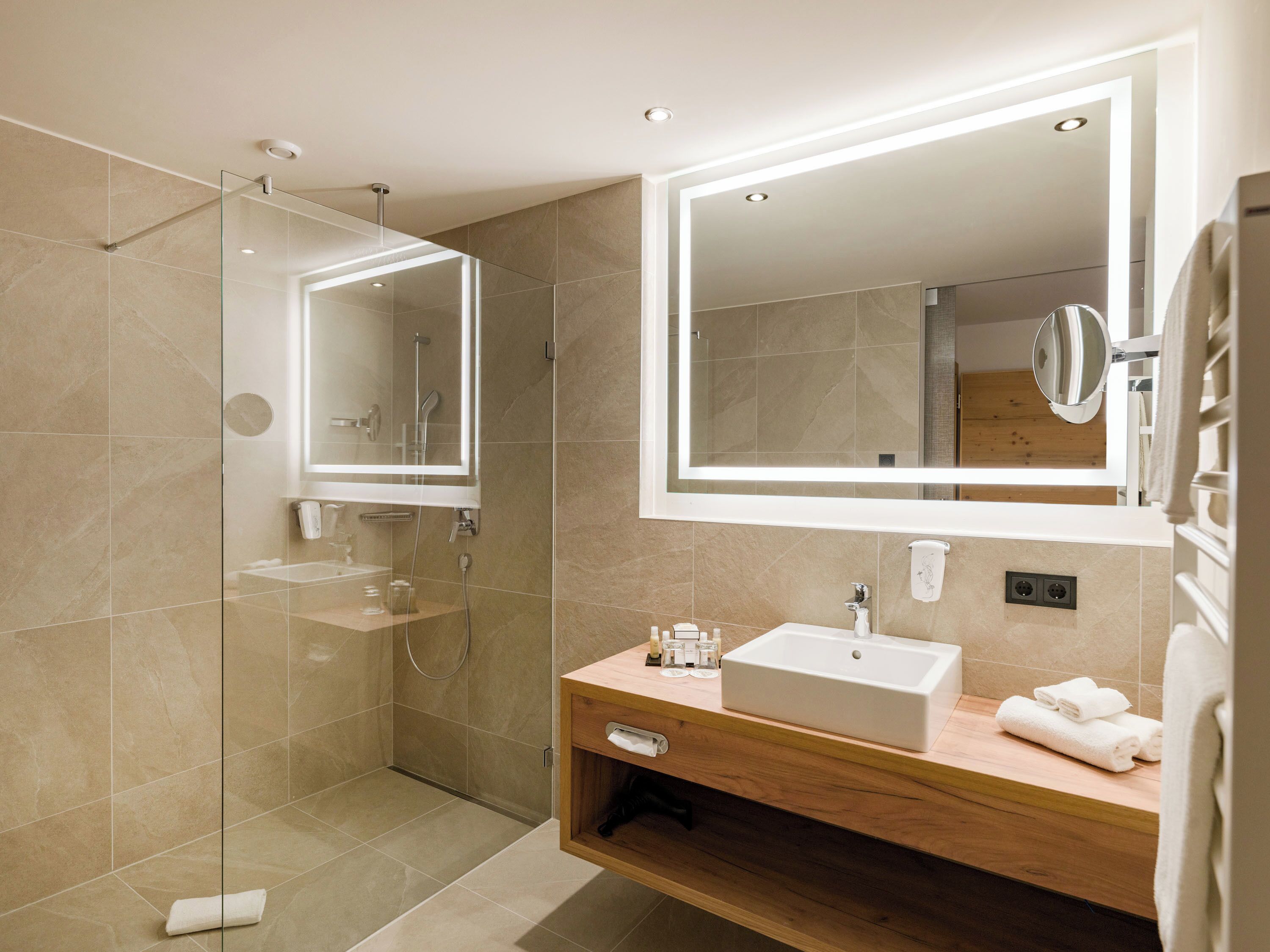 Un baño moderno con ducha envuelta en vidrio, tocador de madera con lavamanos y un espejo grande encima.