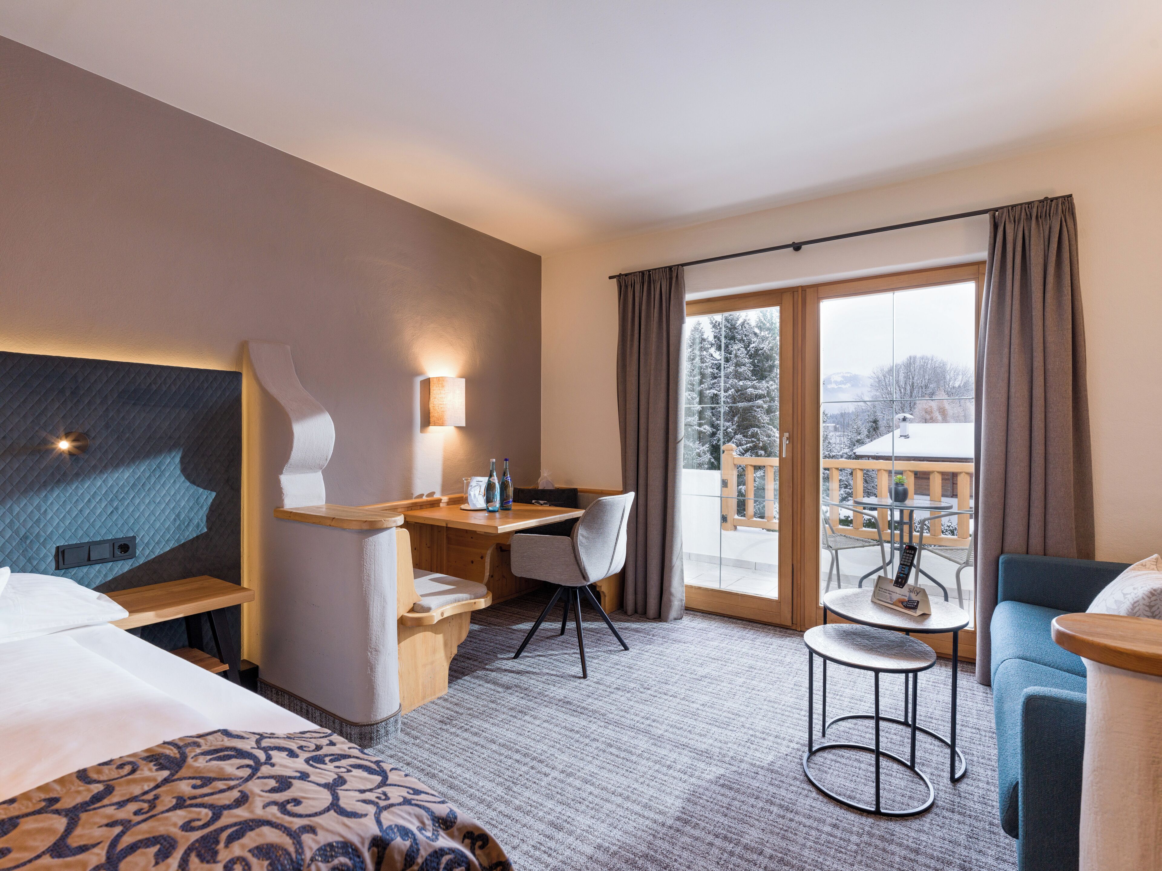 A hotel room with a bed, a desk with a chair, a small table with a chair, and a balcony with a table and chairs.