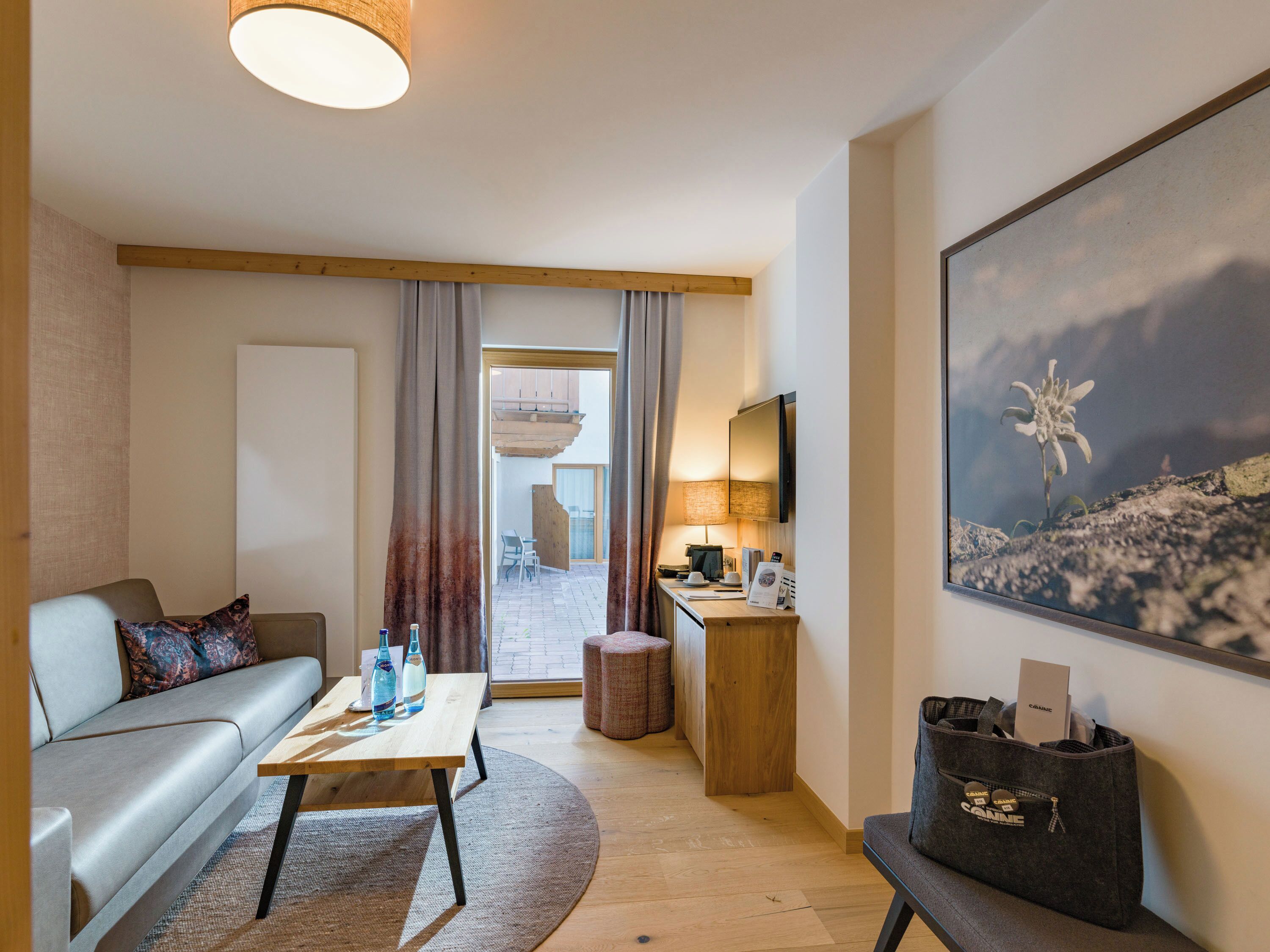 Et moderne hotelværelse med en sofa, et træ sofabord, et skrivebord med lampe, et stort maleri og udsigt til udeområdet.