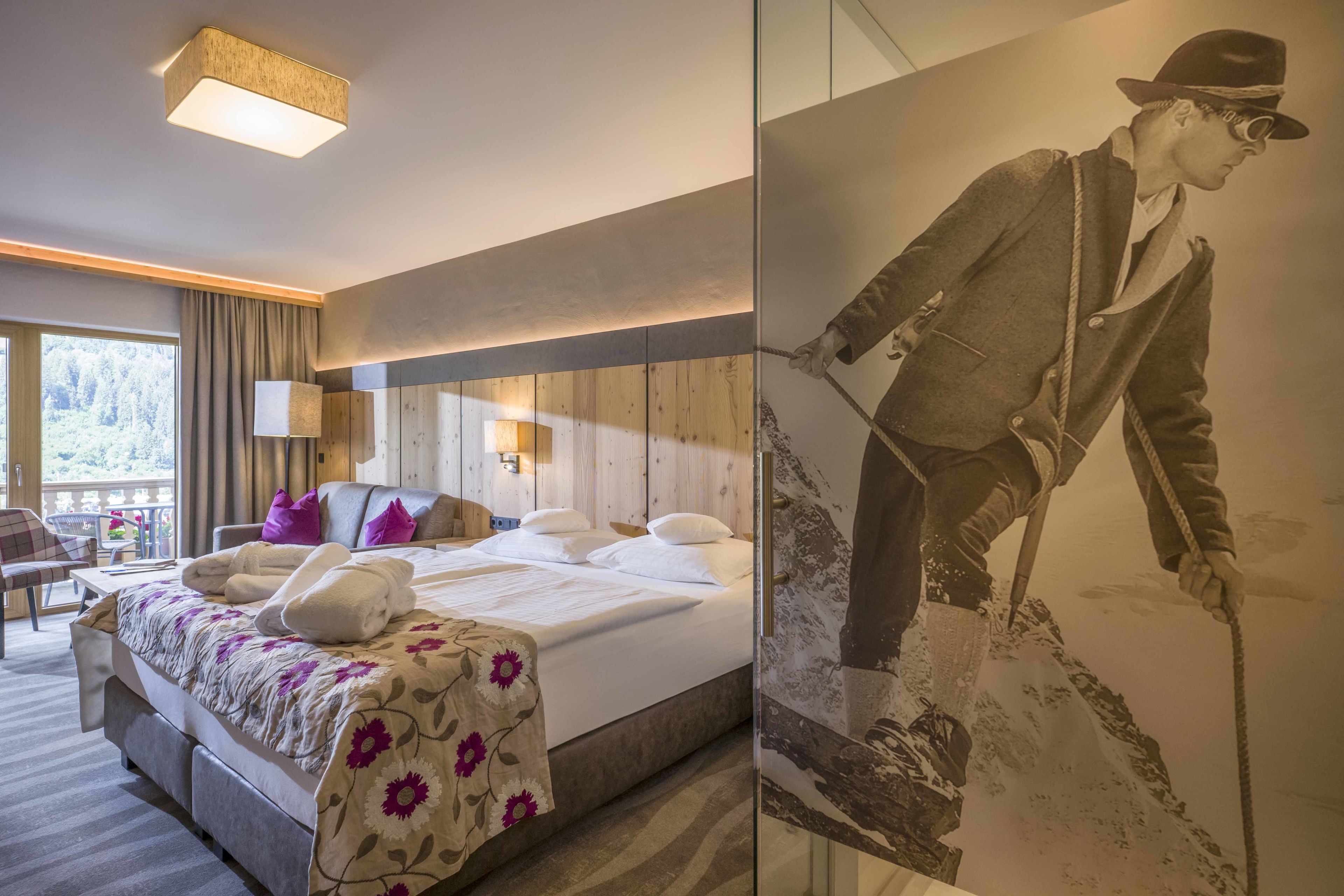 Een hotelkamer met een bed, een bureau en een stoel. Er is een groot raam met gordijnen, een lamp en een schilderij van een man op een berg.