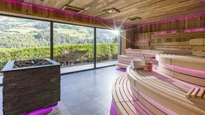 A modern spa area with a stone fireplace, wooden benches, and a large window offering a view of the countryside. -  Hotel Sonne 4 Sterne Superior (Kirchberg in Tirol)