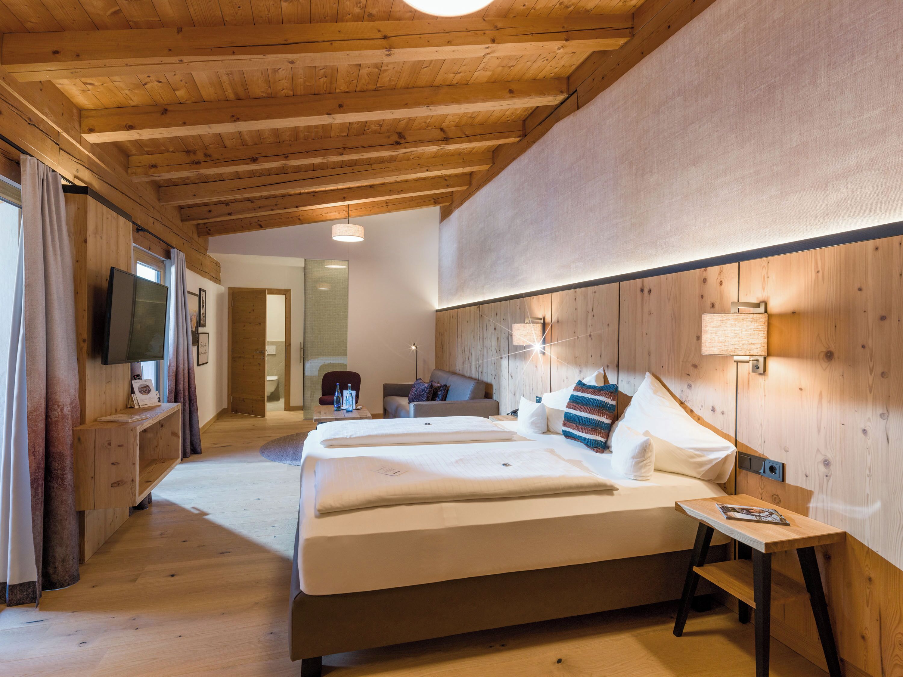 A hotel room with a wooden ceiling, a bed with white bedding, a bedside table, and a TV mounted on the wall.