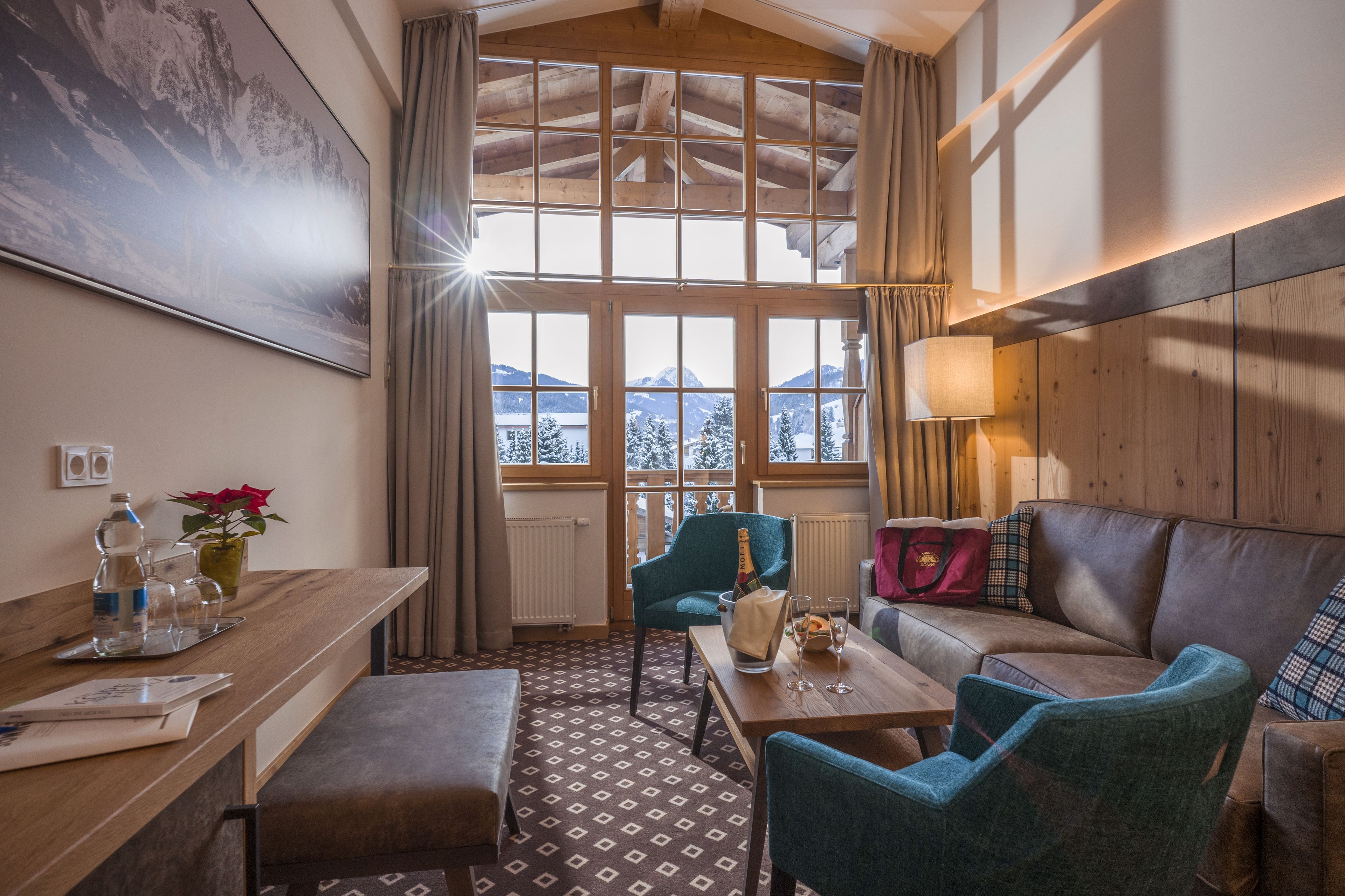 A cozy room with a wooden ceiling, a large window with a mountain view, a wooden table, a blue armchair, and a brown sofa.