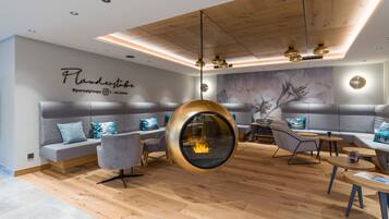 A modern lounge area with a hanging spherical fireplace, grey upholstered seating, and a wooden ceiling.