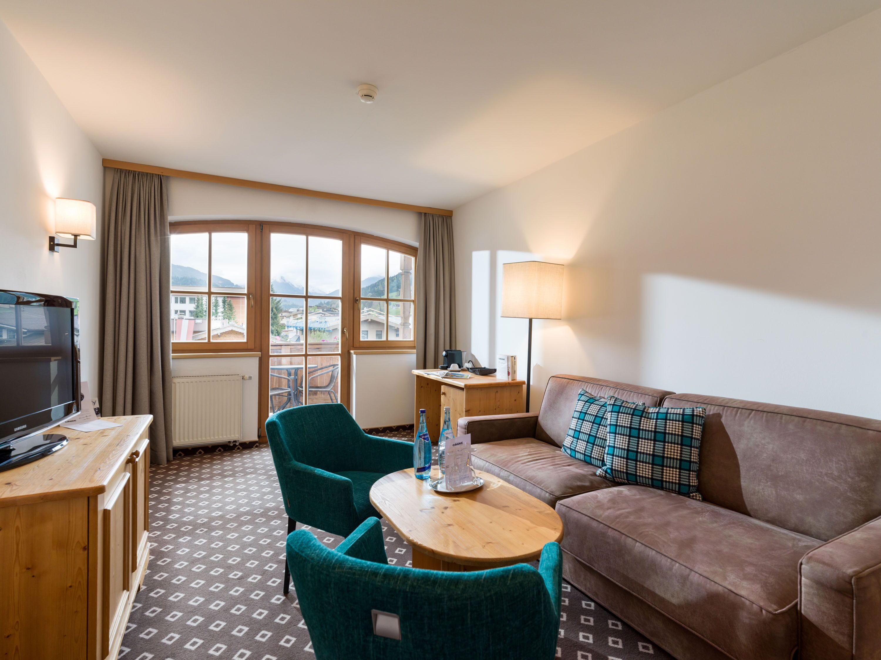 Quarto de hotel com televisão, escrivaninha de madeira, sofá marrom, poltrona verde-água, mesa pequena e janela com vista para edifícios e montanhas.