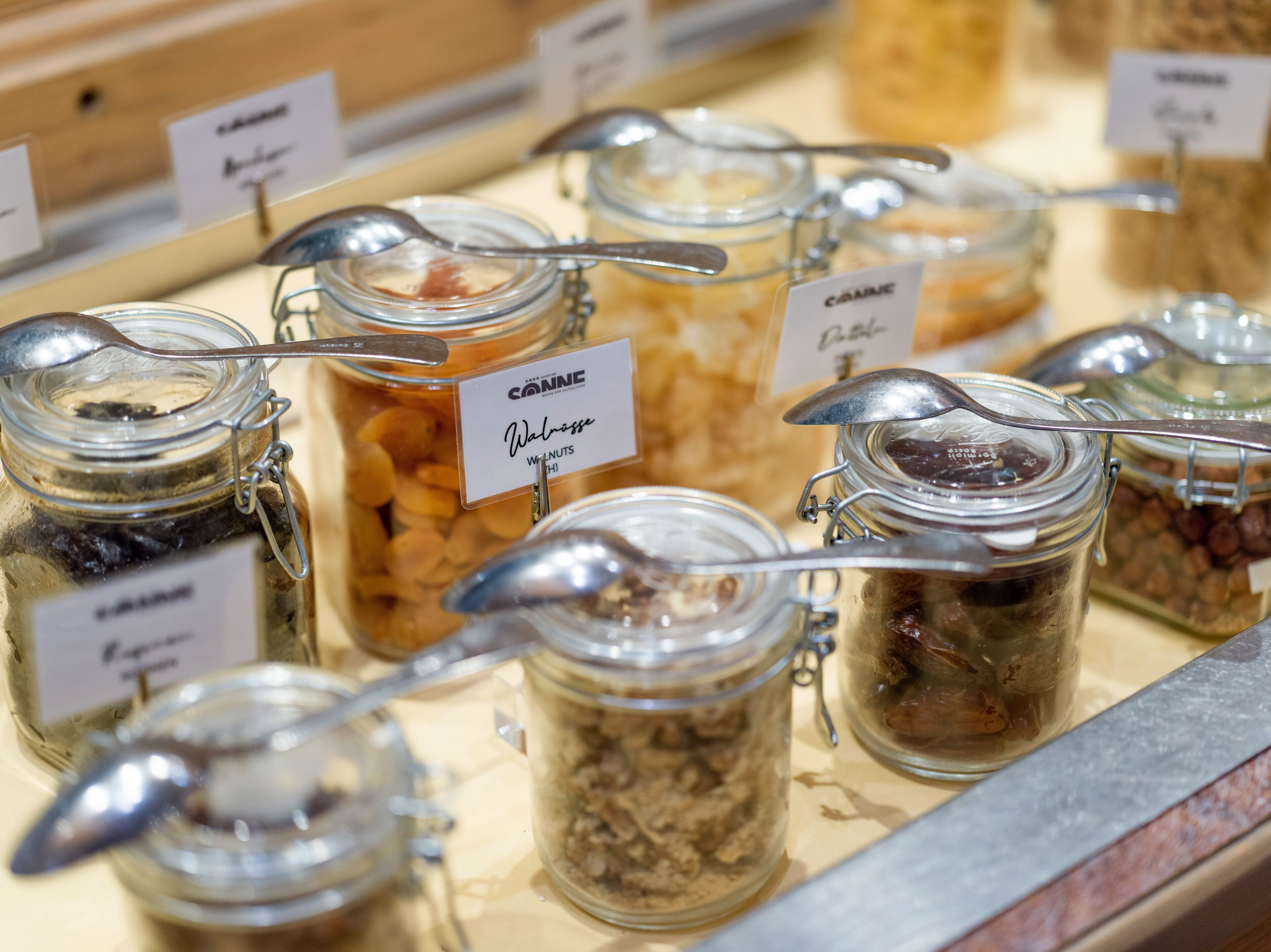 Una presentación de diversos alimentos en frascos de vidrio con tapas metálicas y cucharas, cada uno identificado con el tipo de alimento y la marca.