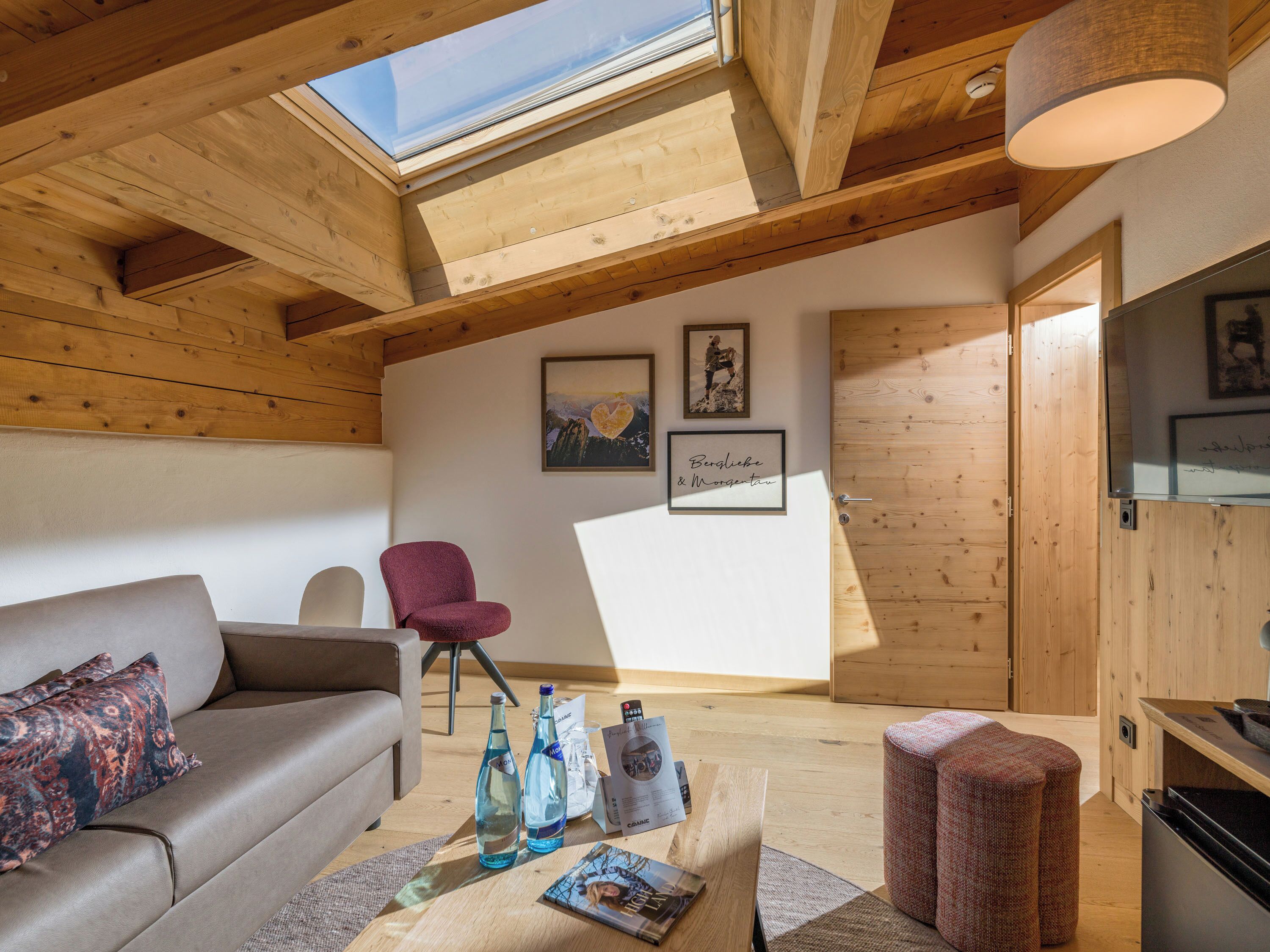 A cozy room with a wooden ceiling, a sofa, a chair, a table with bottles and magazines, and framed pictures on the wall.