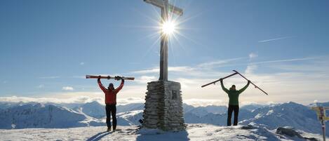 Dos personas sostienen bastones de esquí frente a una cruz en una montaña nevada con cielo despejado.