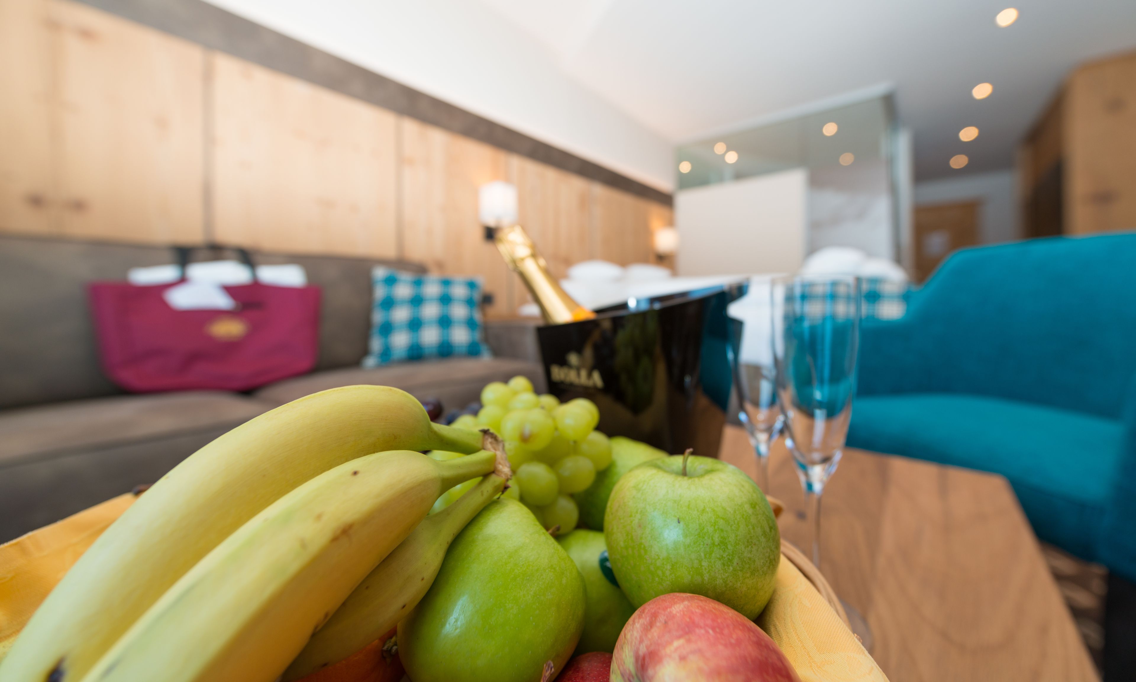 Un frutero con plátanos, uvas y manzanas sobre una mesa de madera en un salón moderno con un sofá azul y paredes de madera.