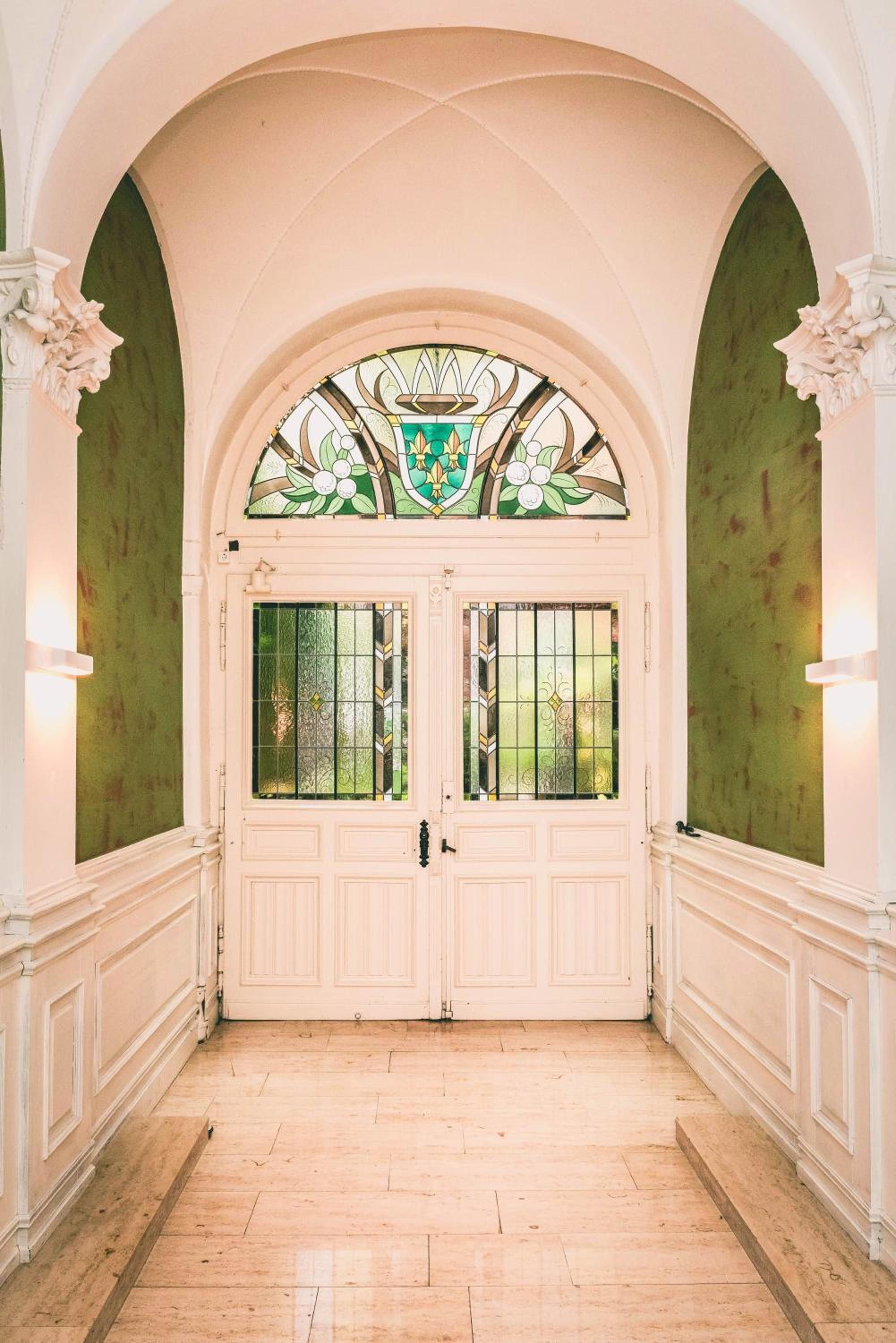 A hallway with a double door, flanked by two columns with decorative capitals, leading to a room with a green wall and a stained glass window above the door.