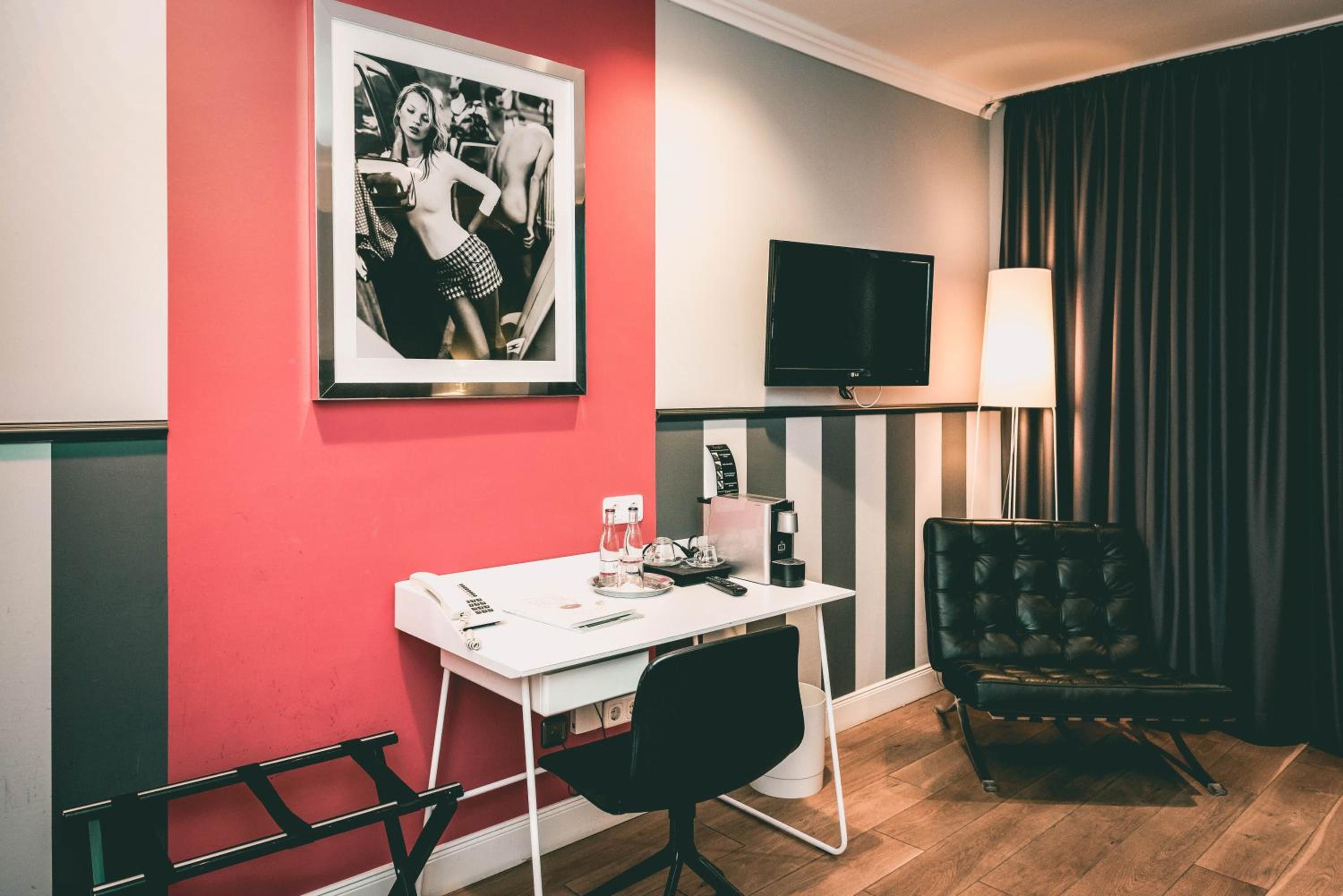 A hotel room with a red accent wall, a framed black and white photo, a flat-screen TV, a desk with a chair, a coffee machine, and a black leather armchair.