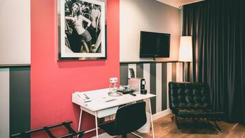 A hotel room with a red accent wall, a framed black and white photo, a flat-screen TV, a desk with a chair, a coffee machine, and a black leather armchair.