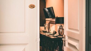 A hallway with a wooden floor leading to a room with a desk, chair, lamp, and a black object on the wall. - Hotel Villa Klemm - Wiesbaden City (Wiesbaden)