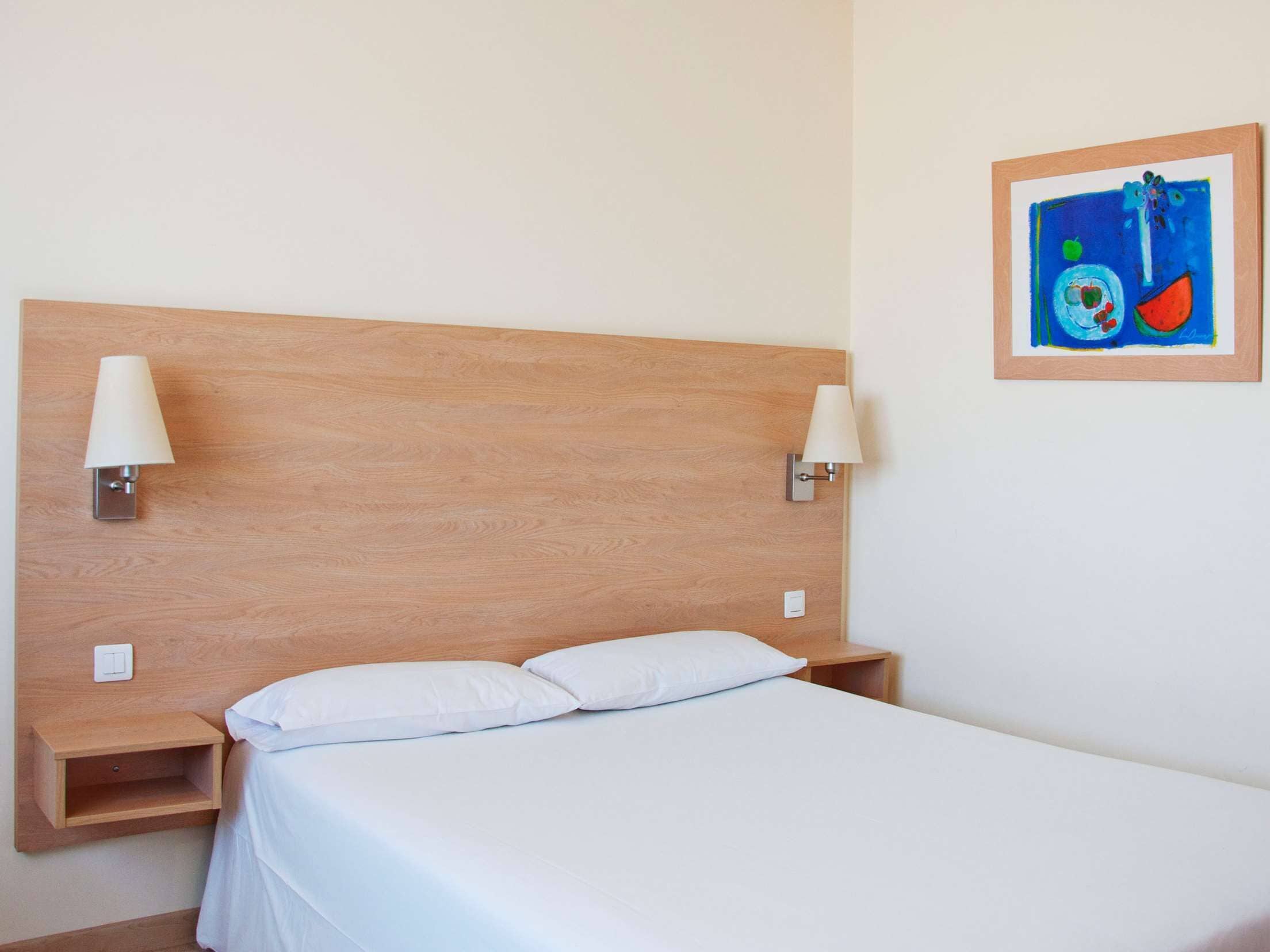 A neatly made bed with white linens, a wooden headboard, and two wall-mounted lamps.