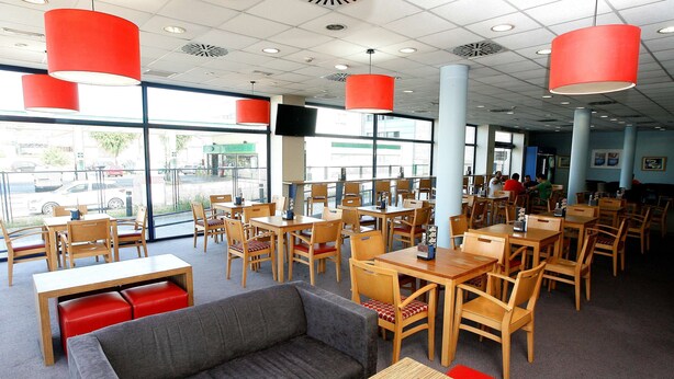 Een ruim restaurant met hoge plafonds, grote ramen en rode hanglampen. Tafels en stoelen zijn klaar voor dineren.