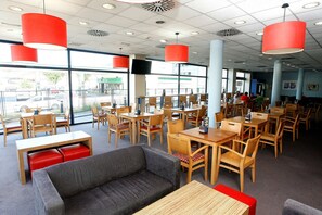 Un restaurant spacieux avec de hauts plafonds, de grandes fenêtres et des lampes suspendues rouges. Les tables et les chaises sont disposées pour accueillir les clients.