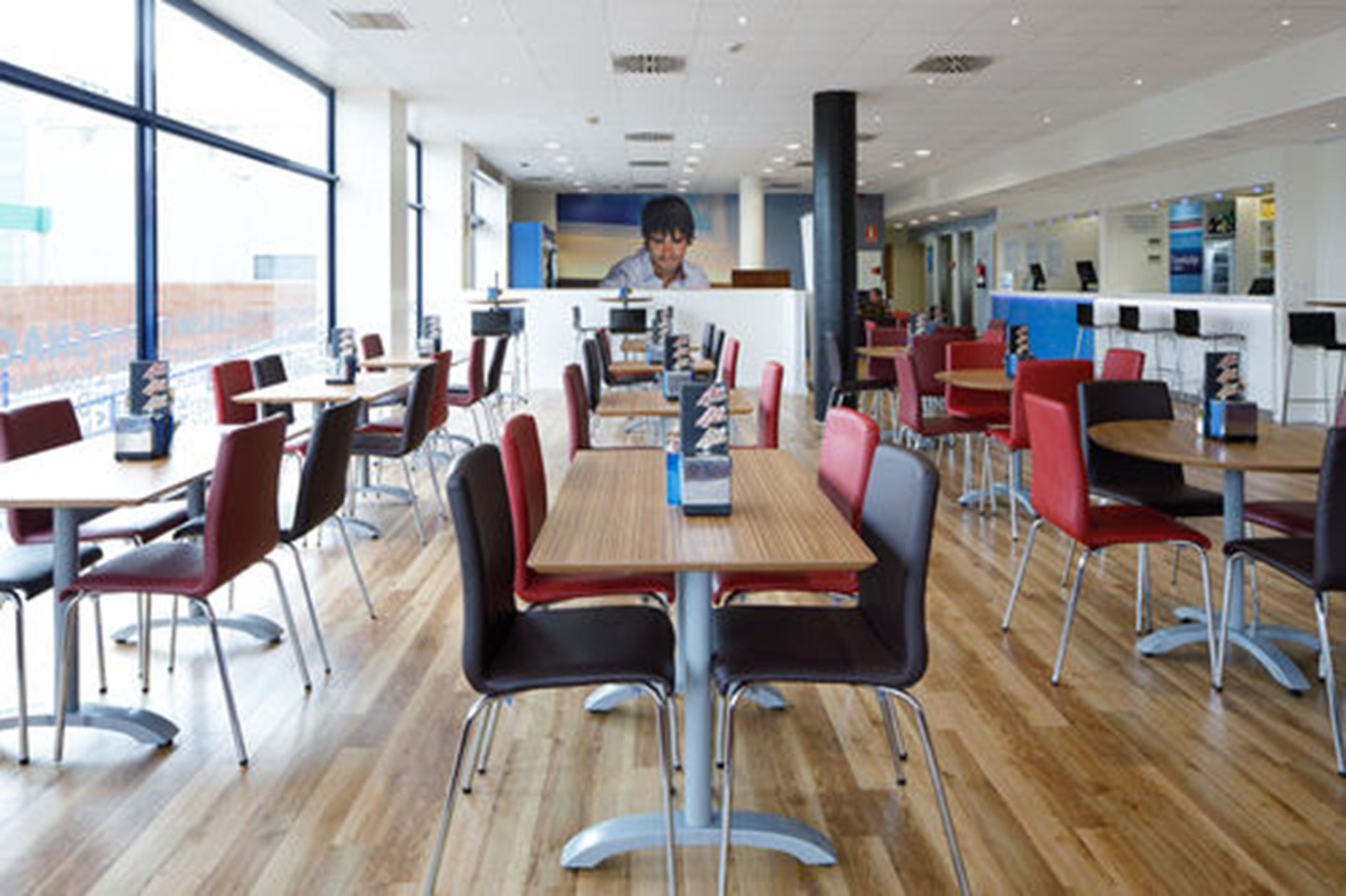 Een moderne cafetaria met houten tafels en stoelen, een bedieningsbalie en grote ramen.