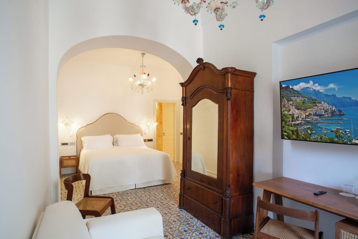 A hotel room with a bed, a wooden wardrobe, a desk with a chair, a television, and a decorative chandelier.