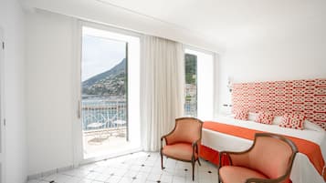 Une chambre d’hôtel comprenant un lit, deux chaises, un balcon avec vue sur la montagne et la mer, et un sol carrelé.
