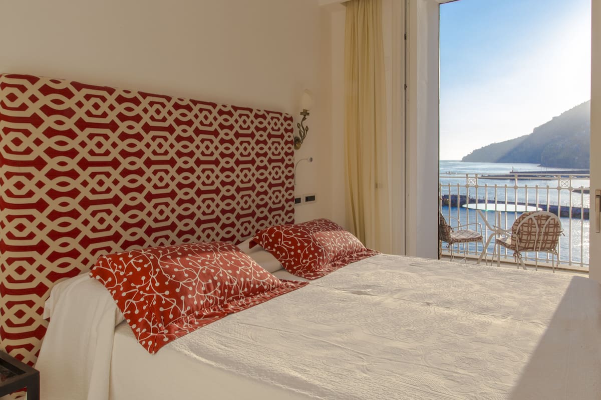 Ein Schlafzimmer mit einem gemusterten Kopfteil, einem Bett mit roter und weißer Bettwäsche und Blick auf einen Balkon mit Stühlen und Geländer.