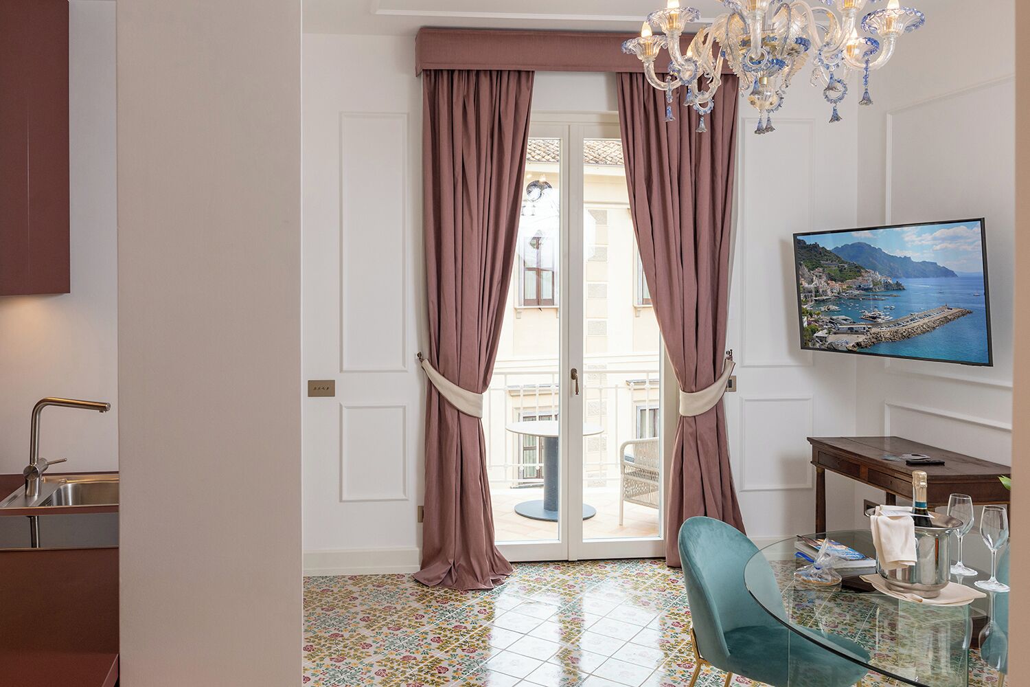 Quarto de hotel com varanda, área de refeições com mesa e cadeiras, TV fixada na parede e um candelabro.