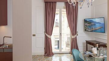 Quarto de hotel com varanda, área de refeições com mesa e cadeiras, TV fixada na parede e um candelabro.