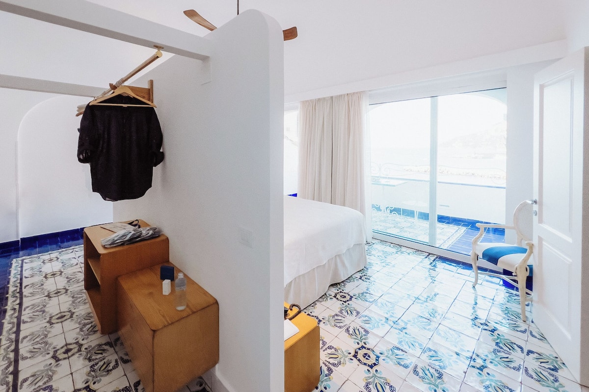 A hotel room with a bed, a small table with a chair, a hanging coat rack, and a large window with a view.