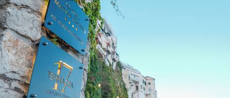 Two signs on a stone wall, one for Hotel Marinaria and the other for Terrazza Bar/Bistro, with a coastal road and buildings in the background.