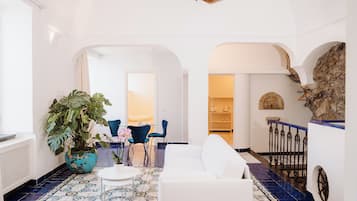 A spacious living area with a white sofa, a coffee table, and a potted plant. There is a blue chair and a dining table in the background. The floor has a patterned tile design.