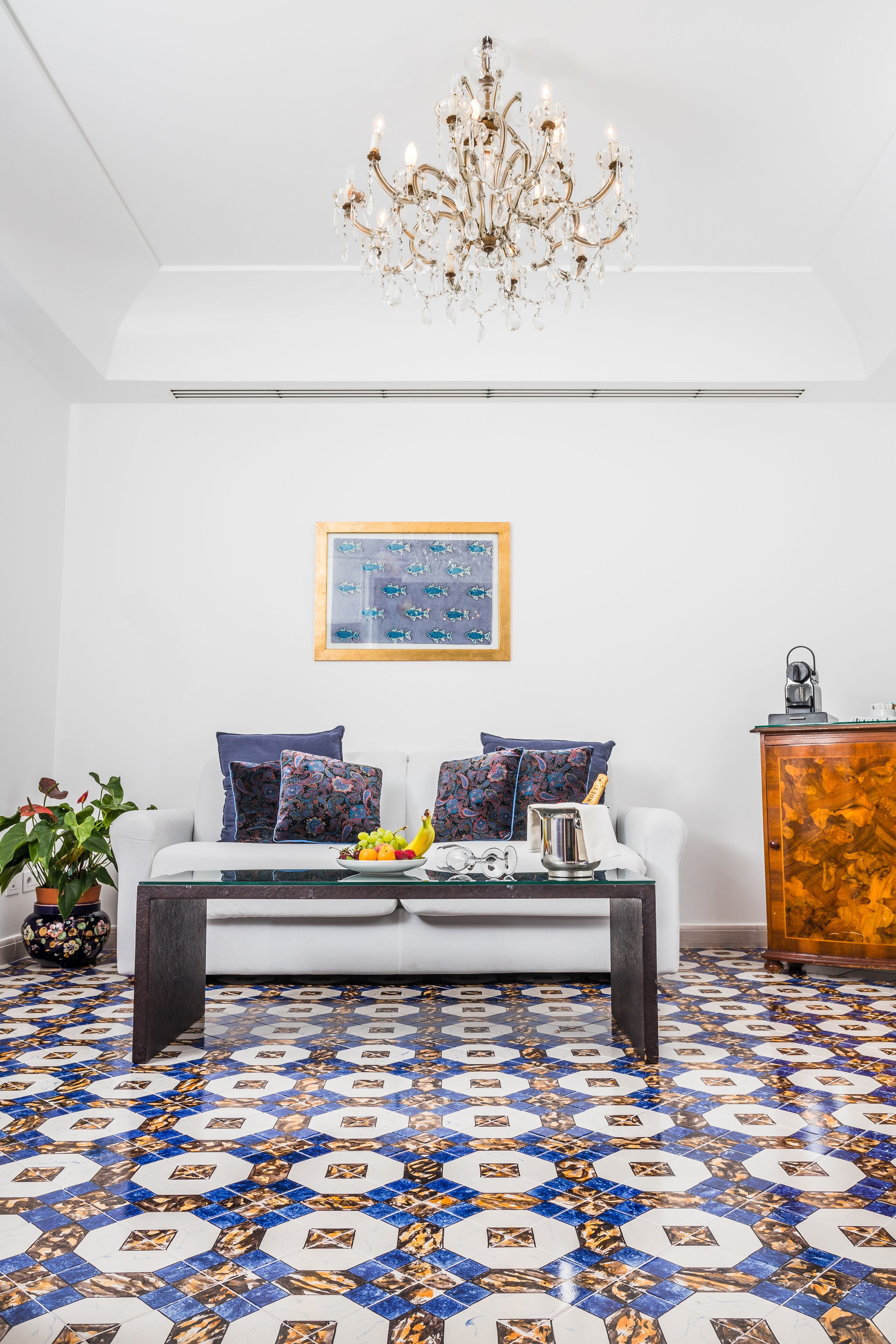 Ein Wohnzimmer mit einem weißen Sofa, gemusterten Kissen, einem Couchtisch, einem Kronleuchter, einem gerahmten Kunstwerk und einem gemusterten Boden.