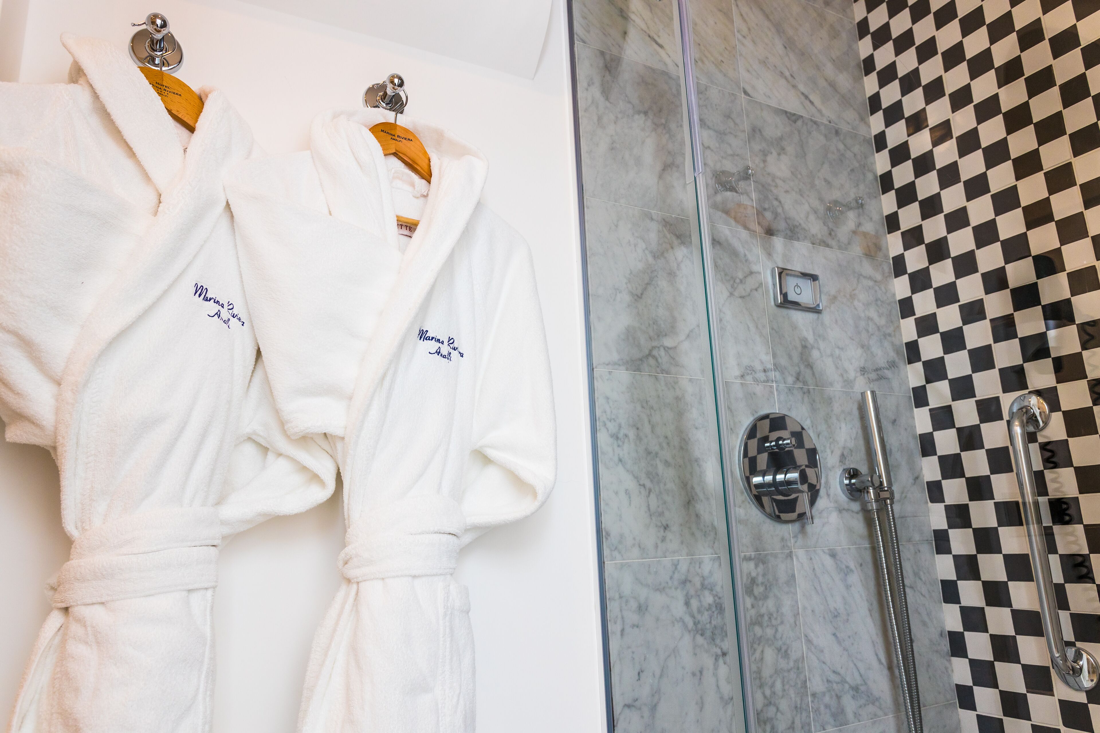 Deux robes blanches ornées de motifs brodés sont accrochées à un mur, une cabine de douche en verre donne sur un espace dont le sol est recouvert de carreaux, et un mur est fait de marbre.