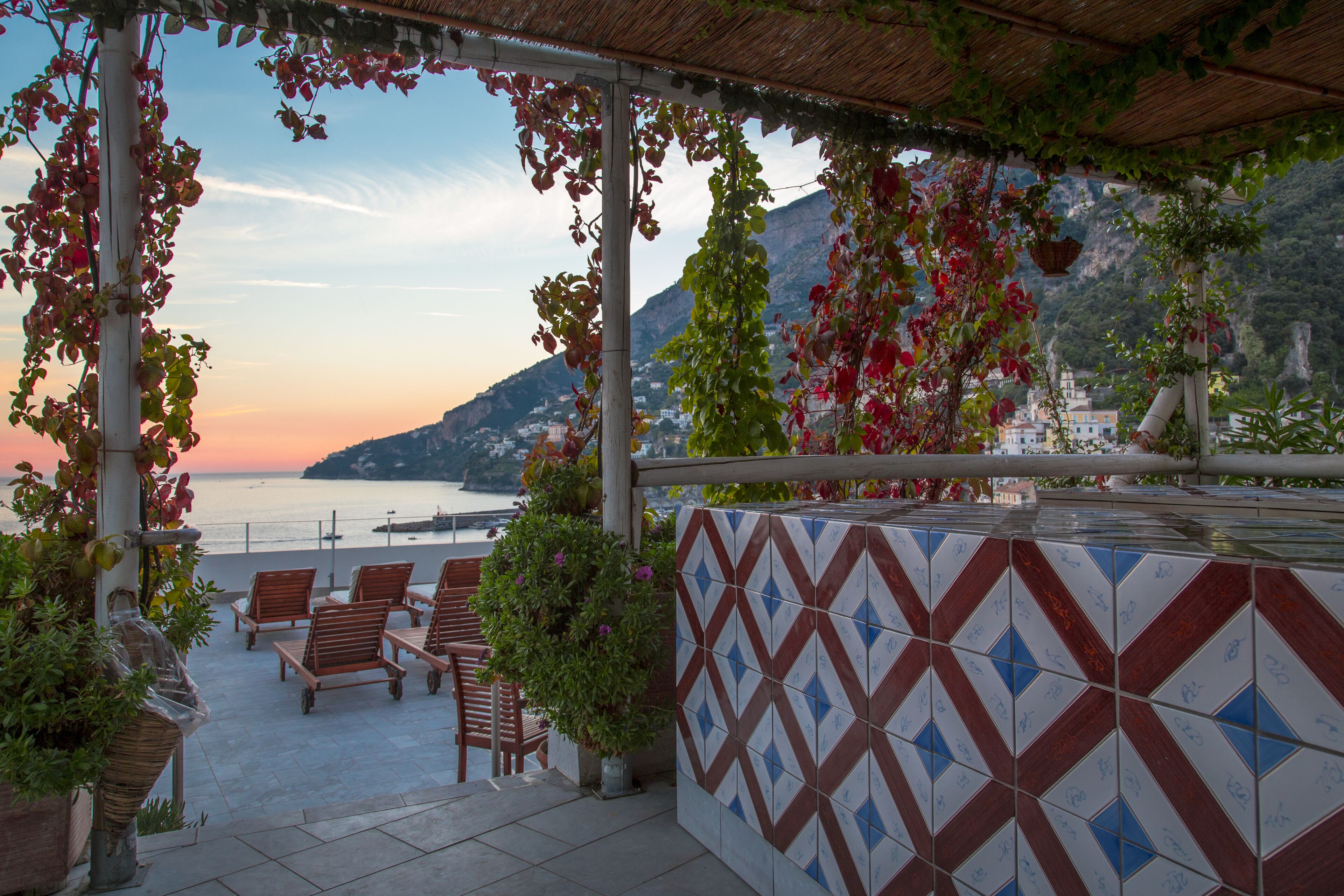 A terrace with chairs and potted plants, overlooking a coastal view with mountains in the background.
