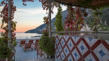 A terrace with chairs and potted plants, overlooking a coastal view with mountains in the background.