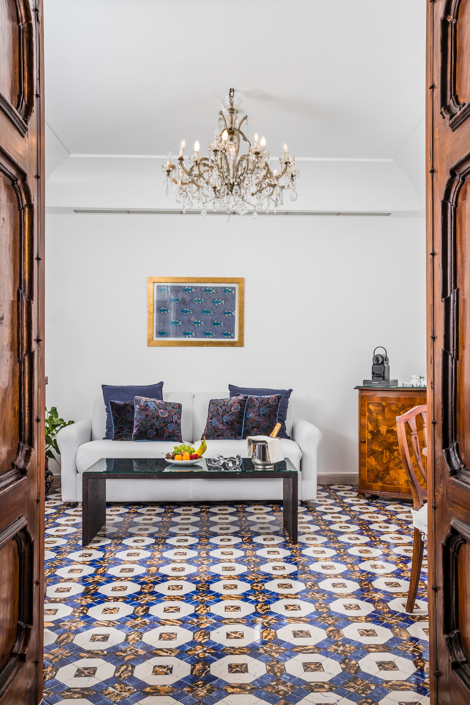 A room with a white sofa, patterned cushions, a glass coffee table, a chandelier, a patterned floor, and a wooden cabinet.