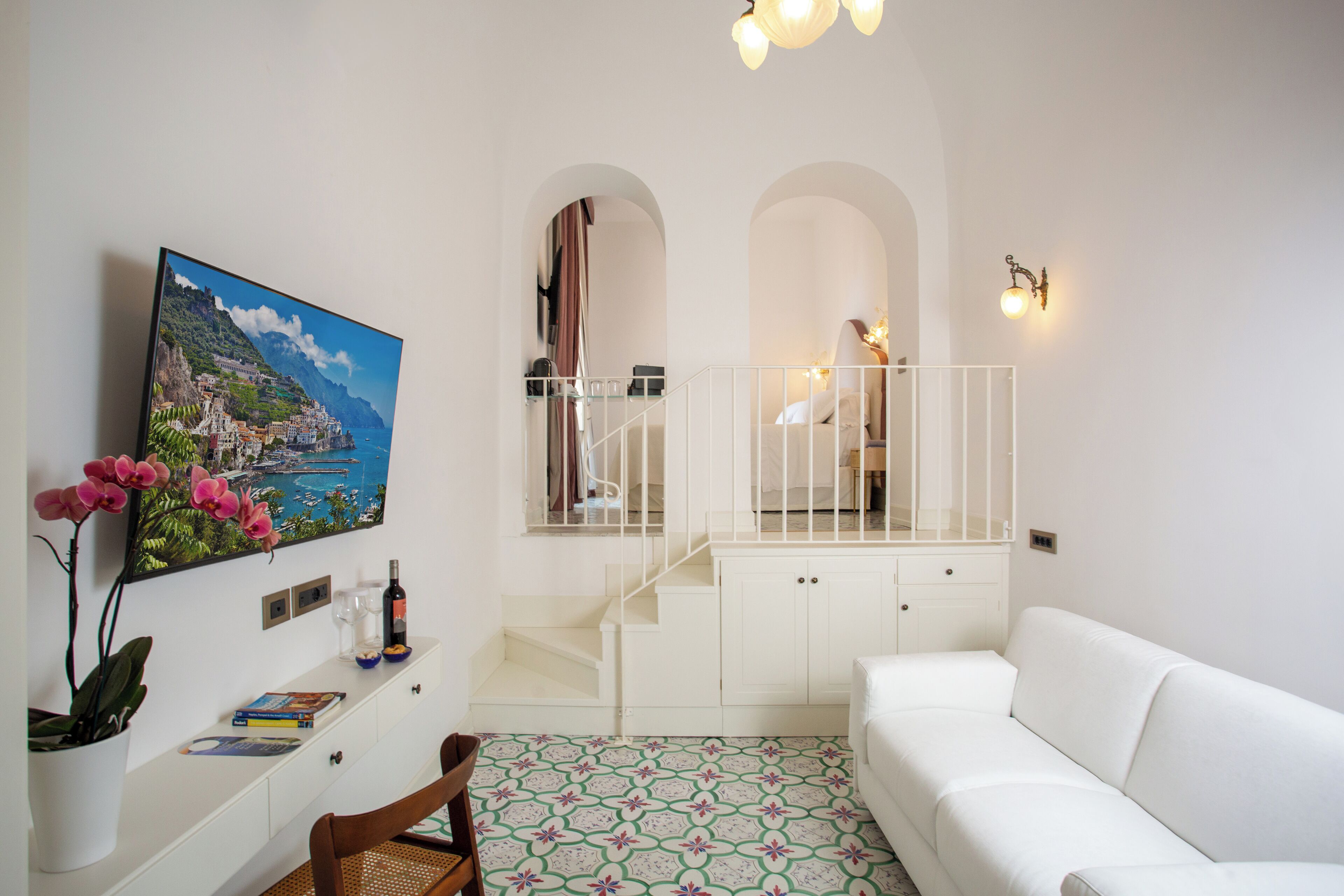 Una sala de estar moderna con un sofá blanco, un televisor montado en la pared, una escalera con barandilla y vista al mar a través de un arco.