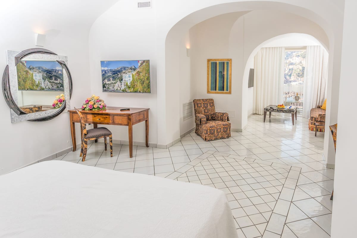 A hotel room with a bed, a desk with a chair, a small table, a mirror, and two framed pictures on the wall.