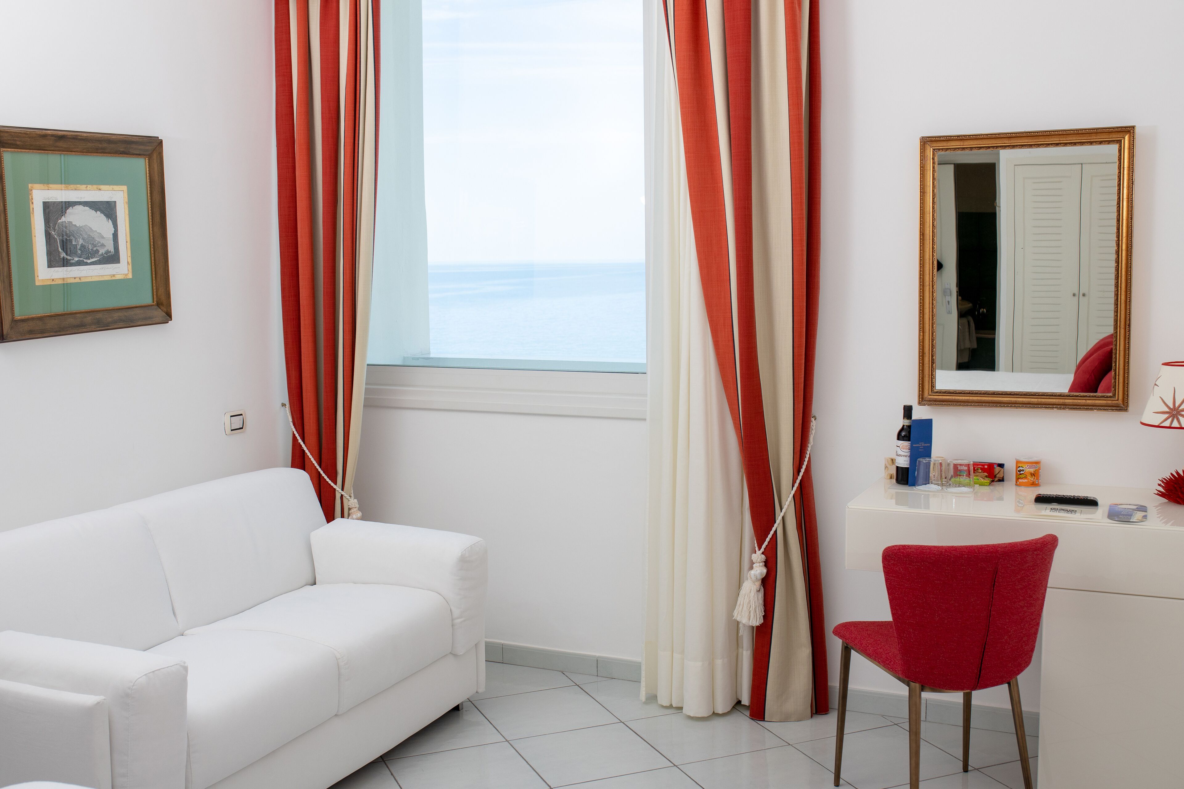 A room with a white sofa, a red chair, a desk with a mirror, and a window with curtains.