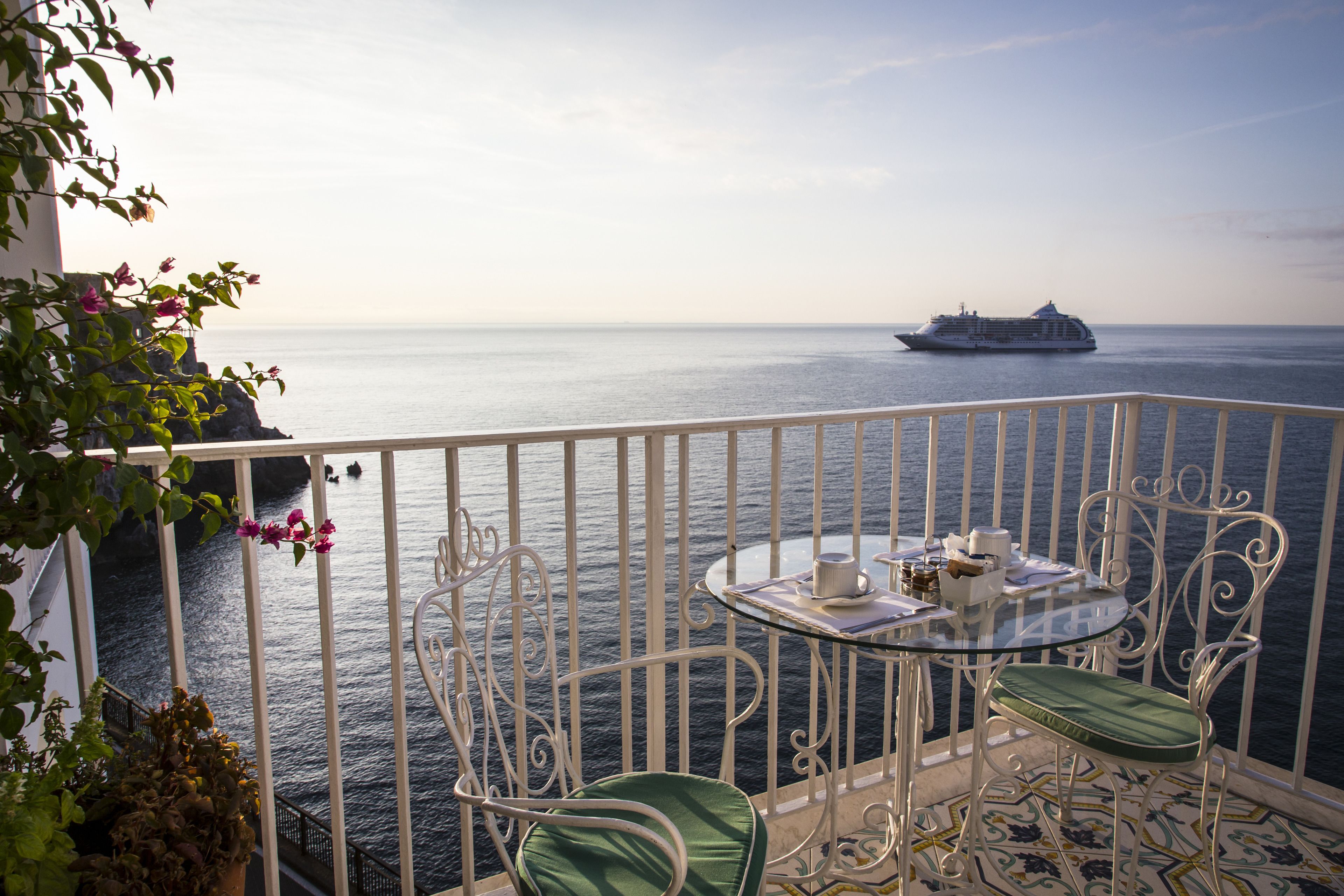 Un balcon avec une table et des chaises, donnant sur une mer calme où l’on aperçoit, au loin, un navire de croisière.