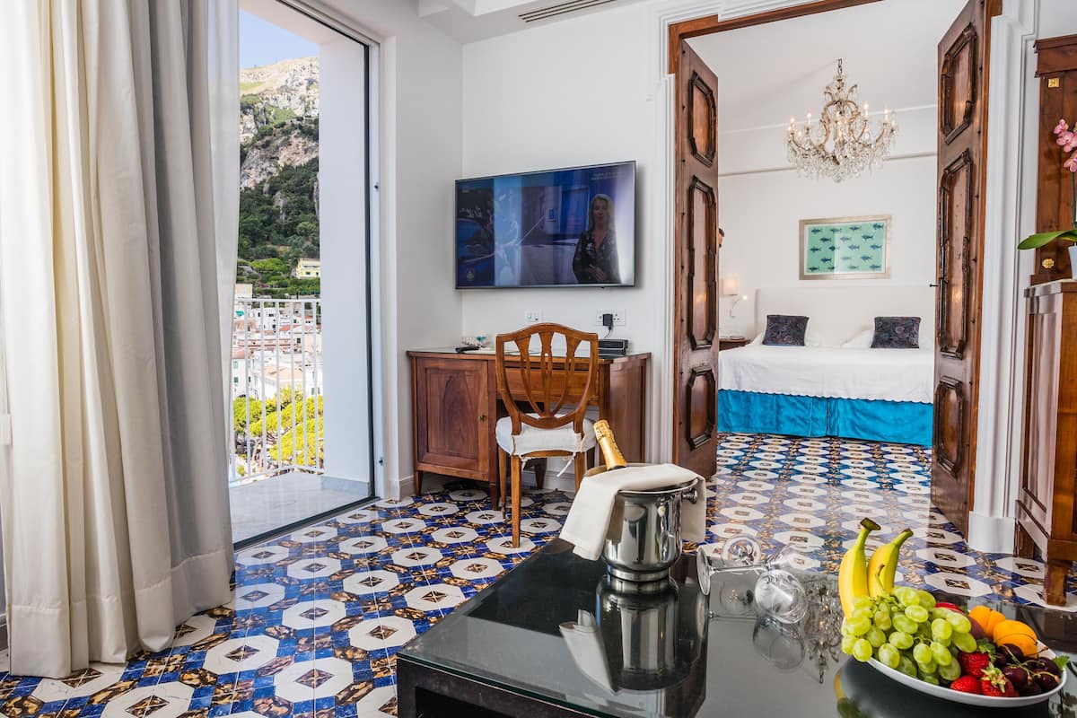 A hotel room with a balcony, a television, a bed with a blue and white bedspread, and a table with fruit and a coffee pot.