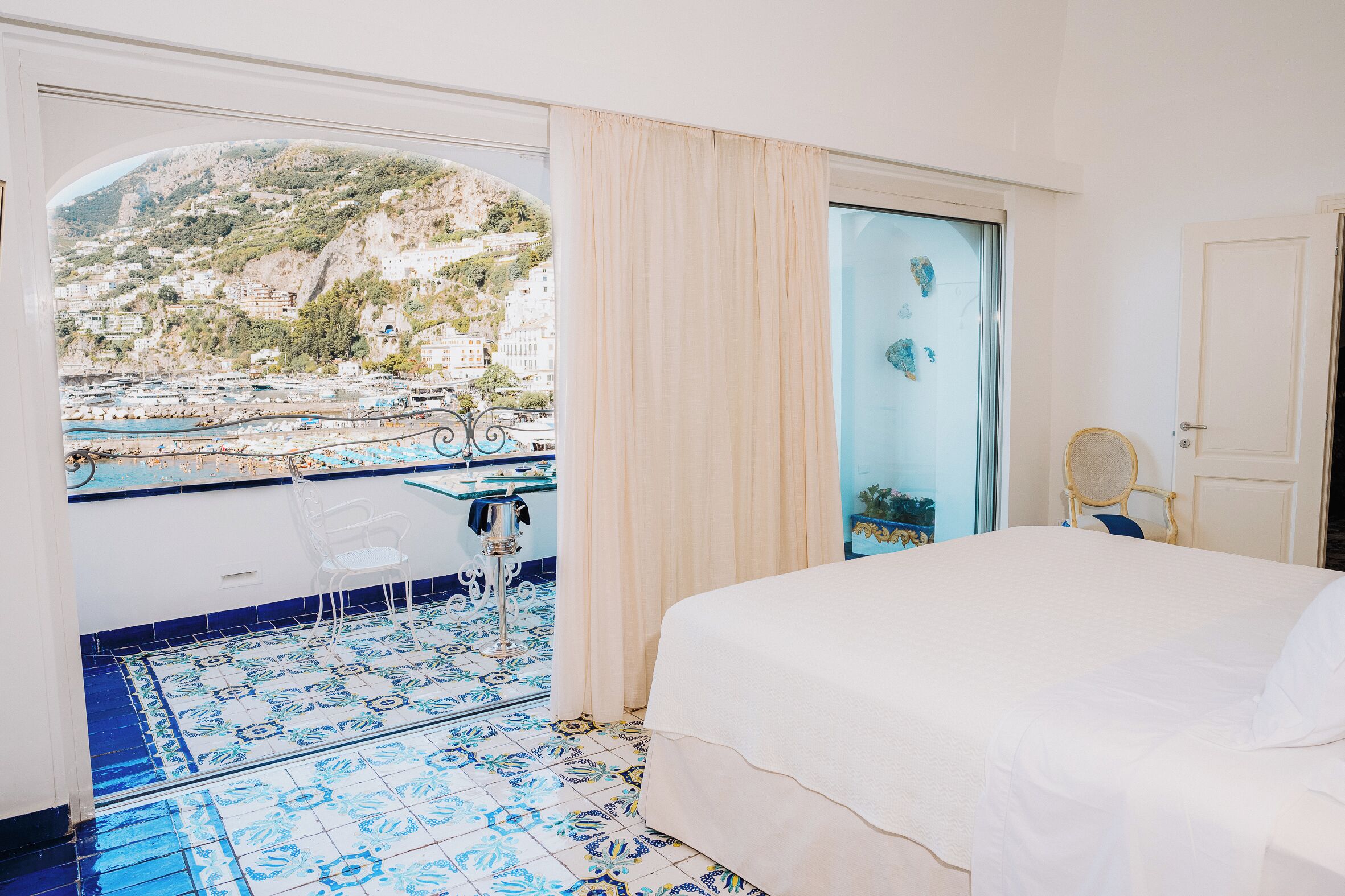 A hotel room with a bed, a chair, a table, and a view of a coastal town.