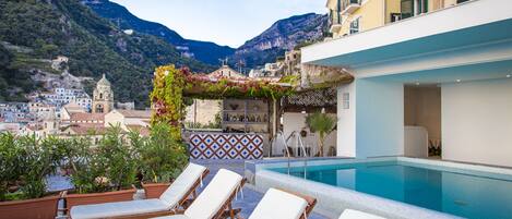 A rooftop pool area with lounge chairs, a bar, and a view of a town and mountains.
