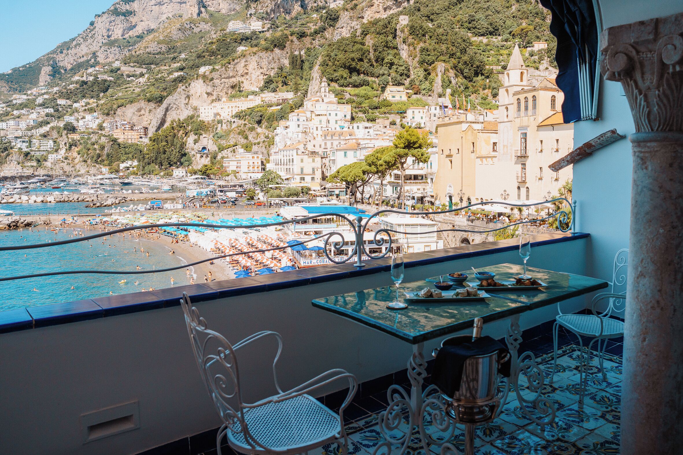 A balcony with a table, chairs, and a view of a coastal town with buildings and a beach.